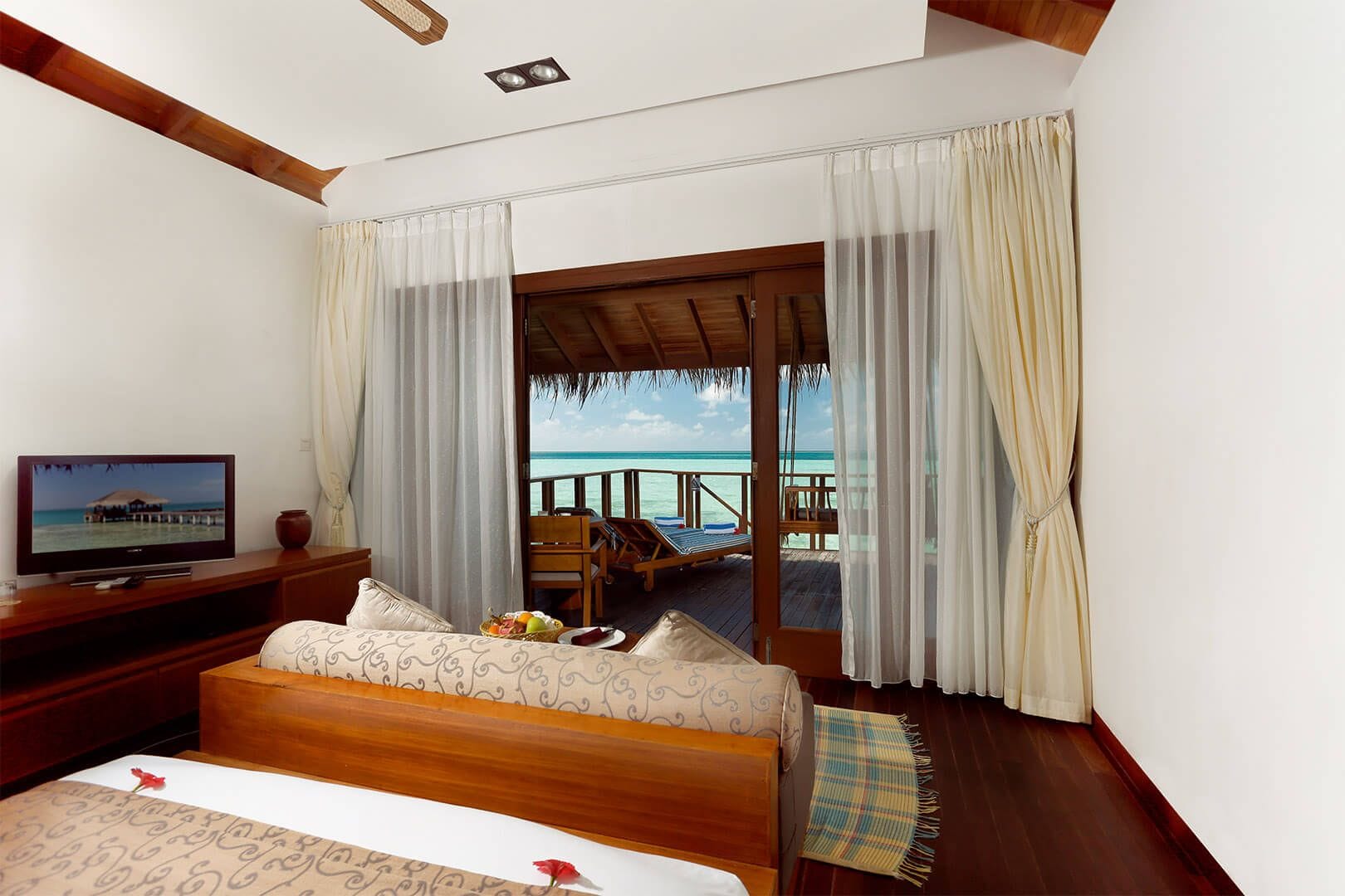 Quarto de hotel com cama, televisão, vista para o oceano e varanda com deck de madeira.