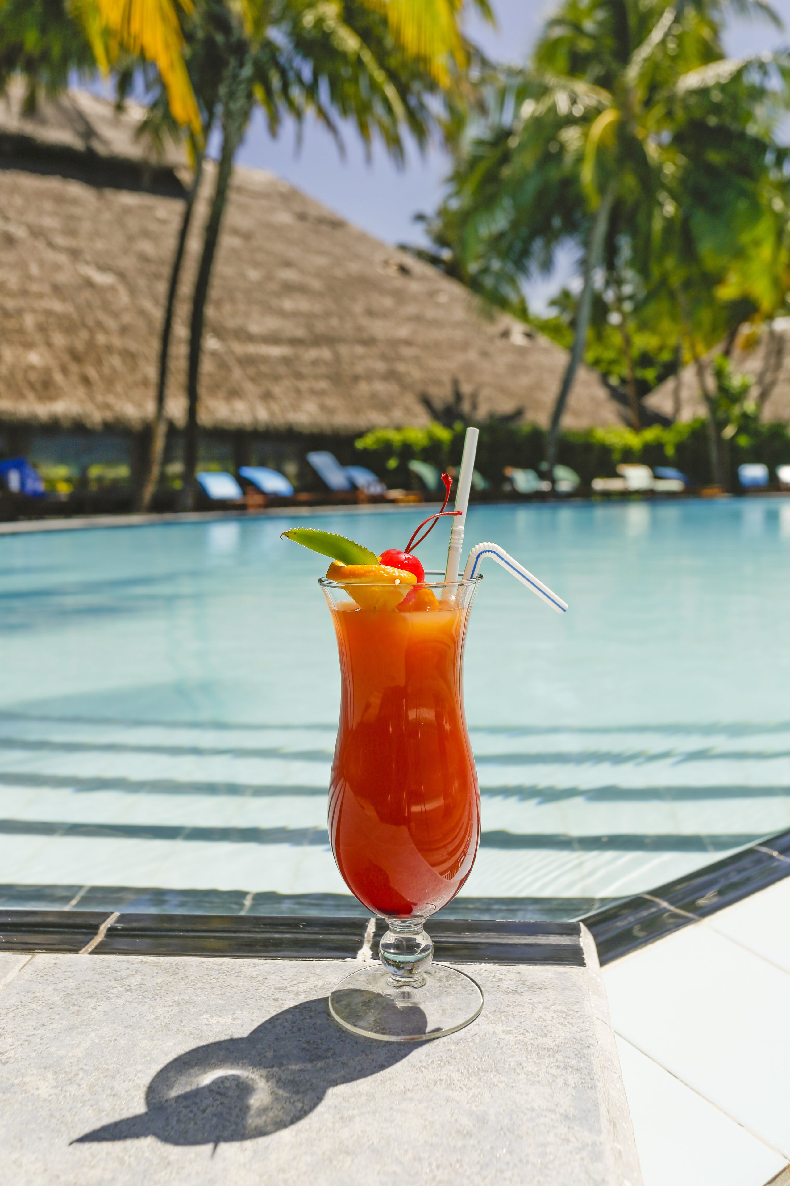 A tall glass with a red beverage, garnished with a cherry and a mint leaf, placed on a tiled surface by a pool with lounge chairs and palm trees in the background.