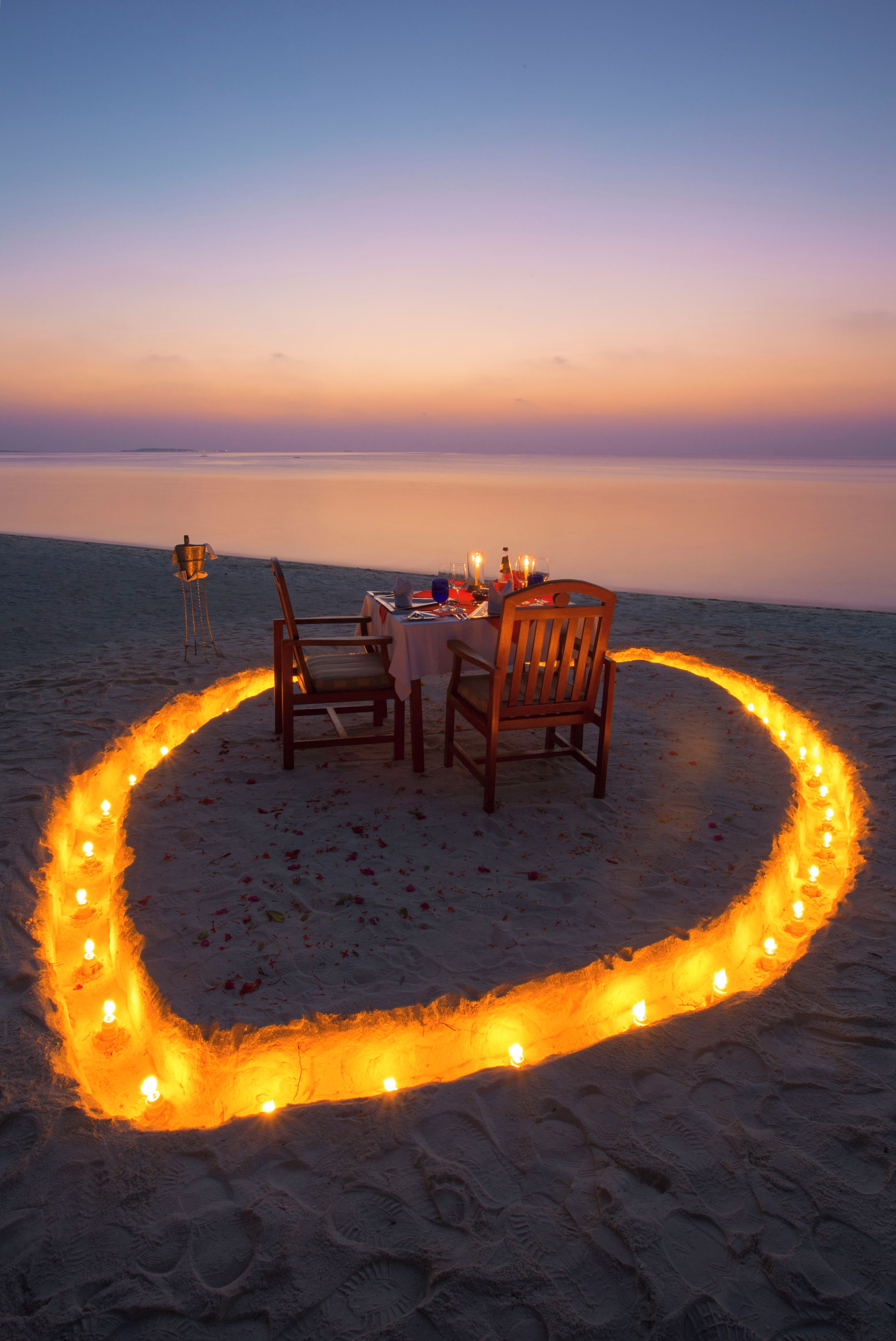 Un montaje para comer en la playa con una mesa y sillas dispuestas sobre la arena, rodeado por un círculo de velas.