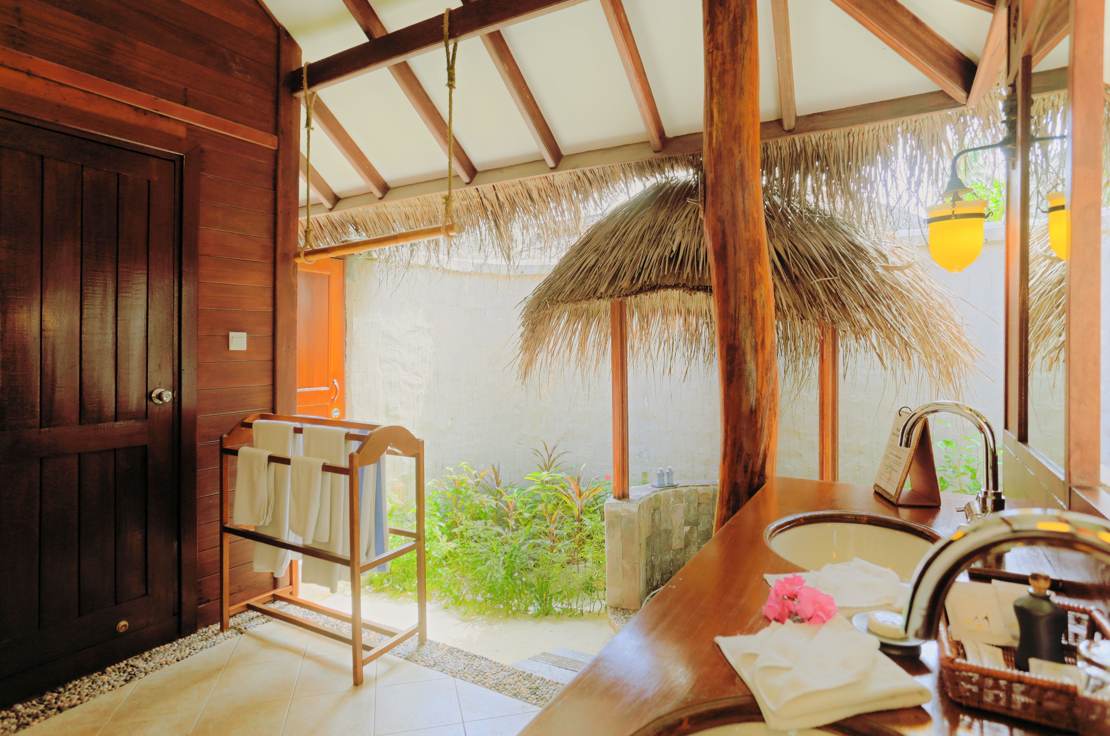 A wooden interior with a thatched roof, a wooden door, a towel rack, a sink with toiletries, and a view of greenery outside.
