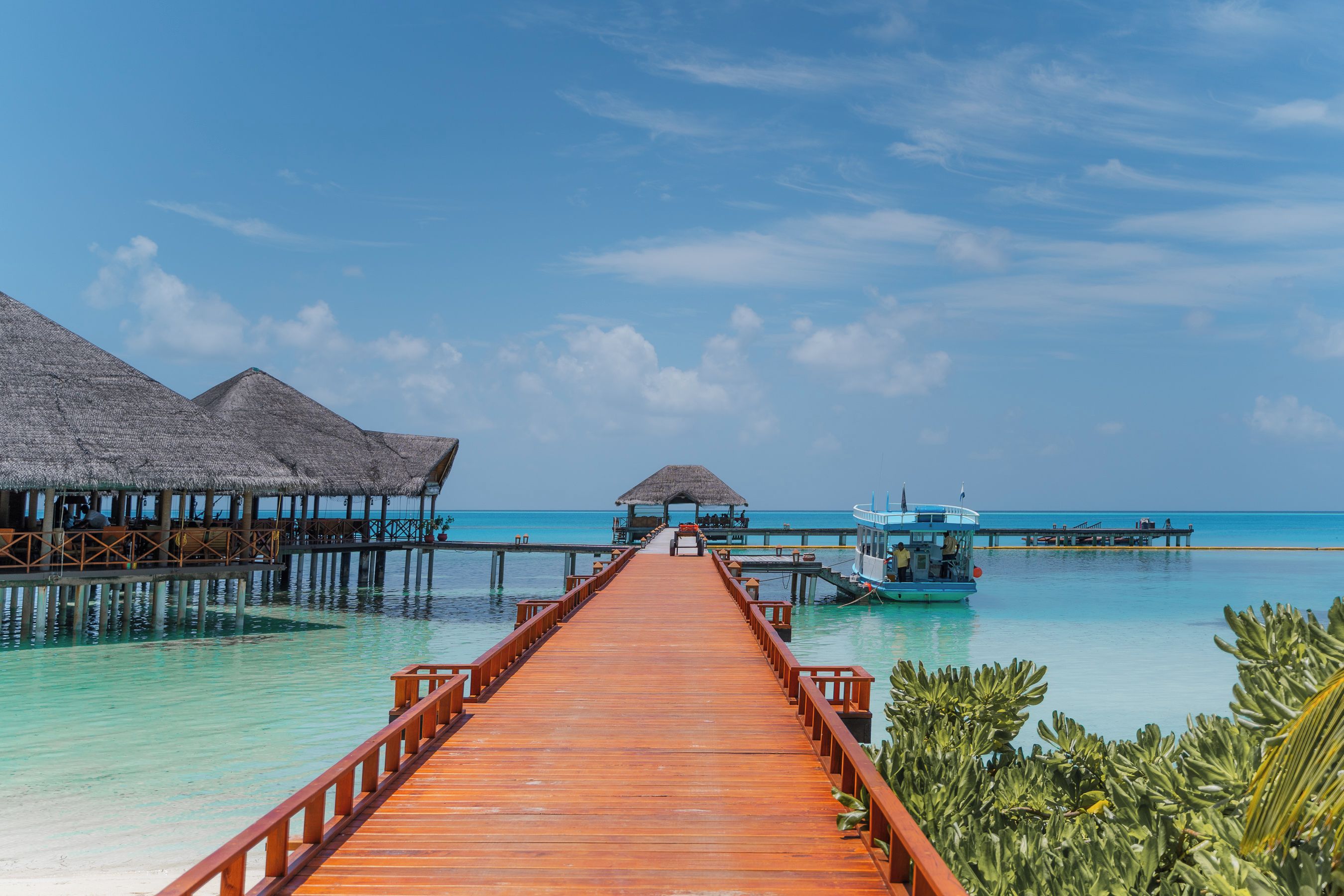 A wooden pier leading to overwater bungalows with thatched roofs, a boat docked at the end, and clear turquoise waters.