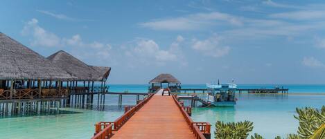 A wooden pier leading to overwater bungalows with thatched roofs, a boat docked at the end, and clear turquoise waters.