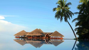 Overwater bungalows with thatched roofs, a clear blue sky, and a calm water surface reflecting the structures.
