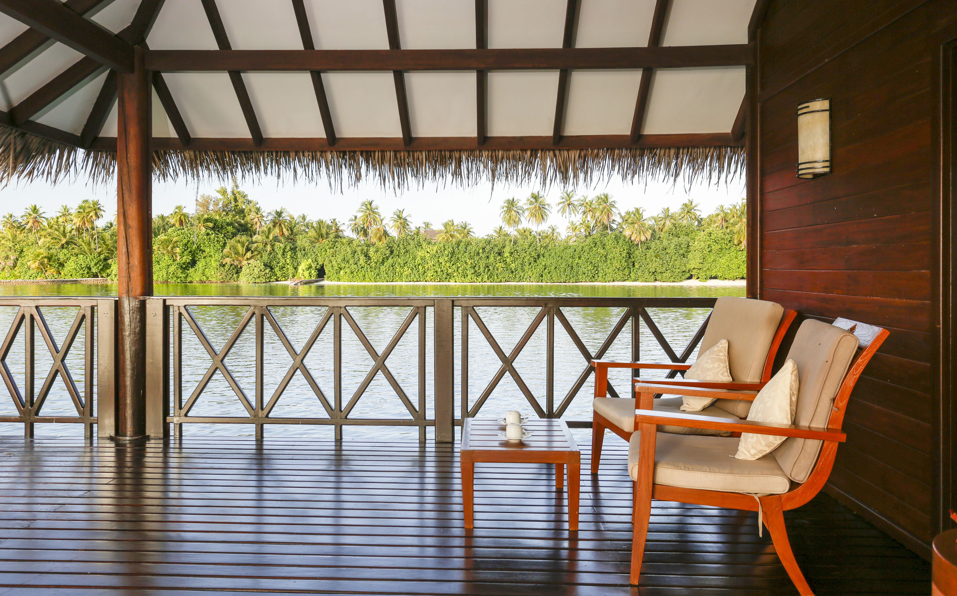 A wooden deck with a railing, a table, and two chairs with cushions, overlooking a body of water and greenery.
