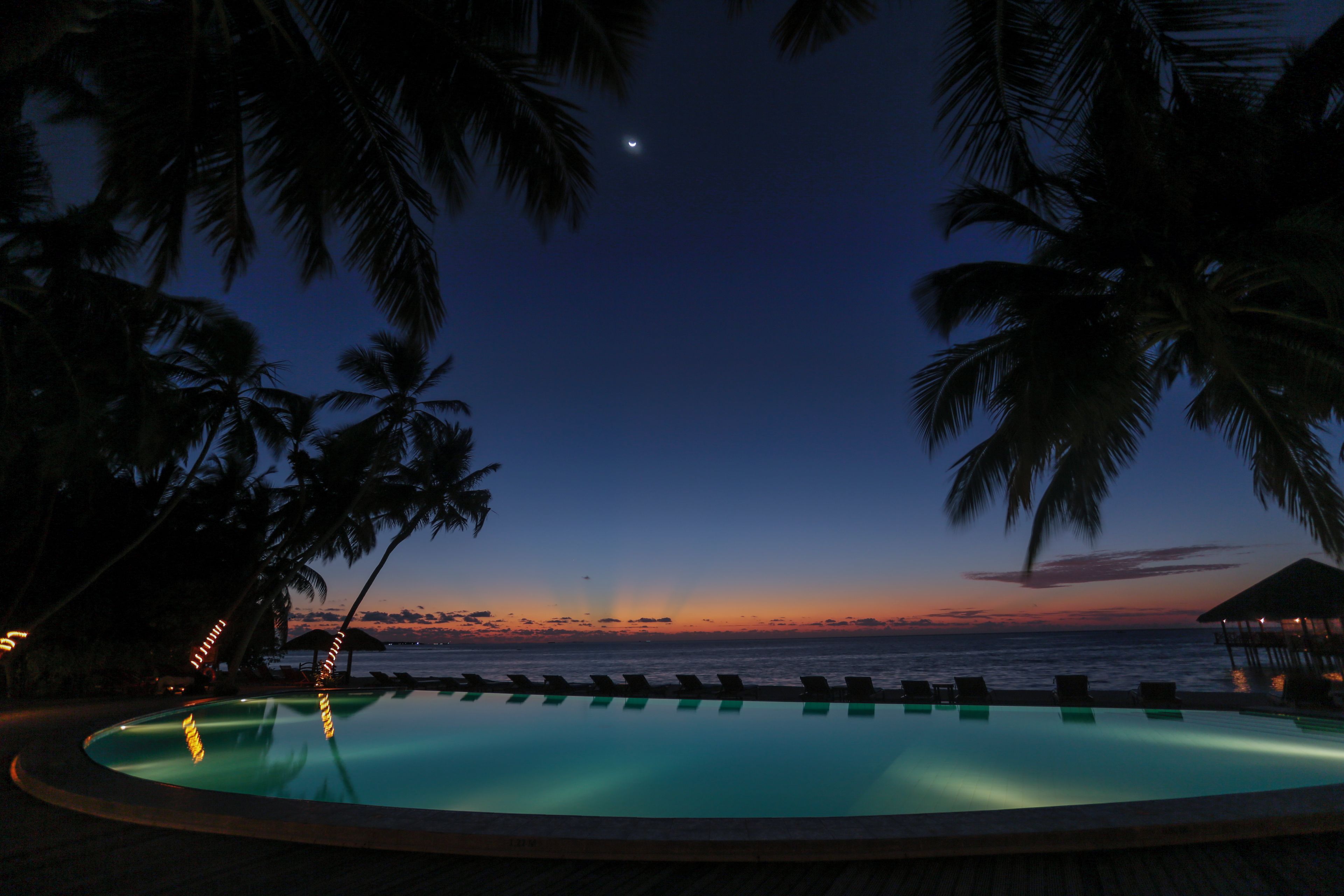 Una piscina con vistas al océano al atardecer, rodeada de palmeras.