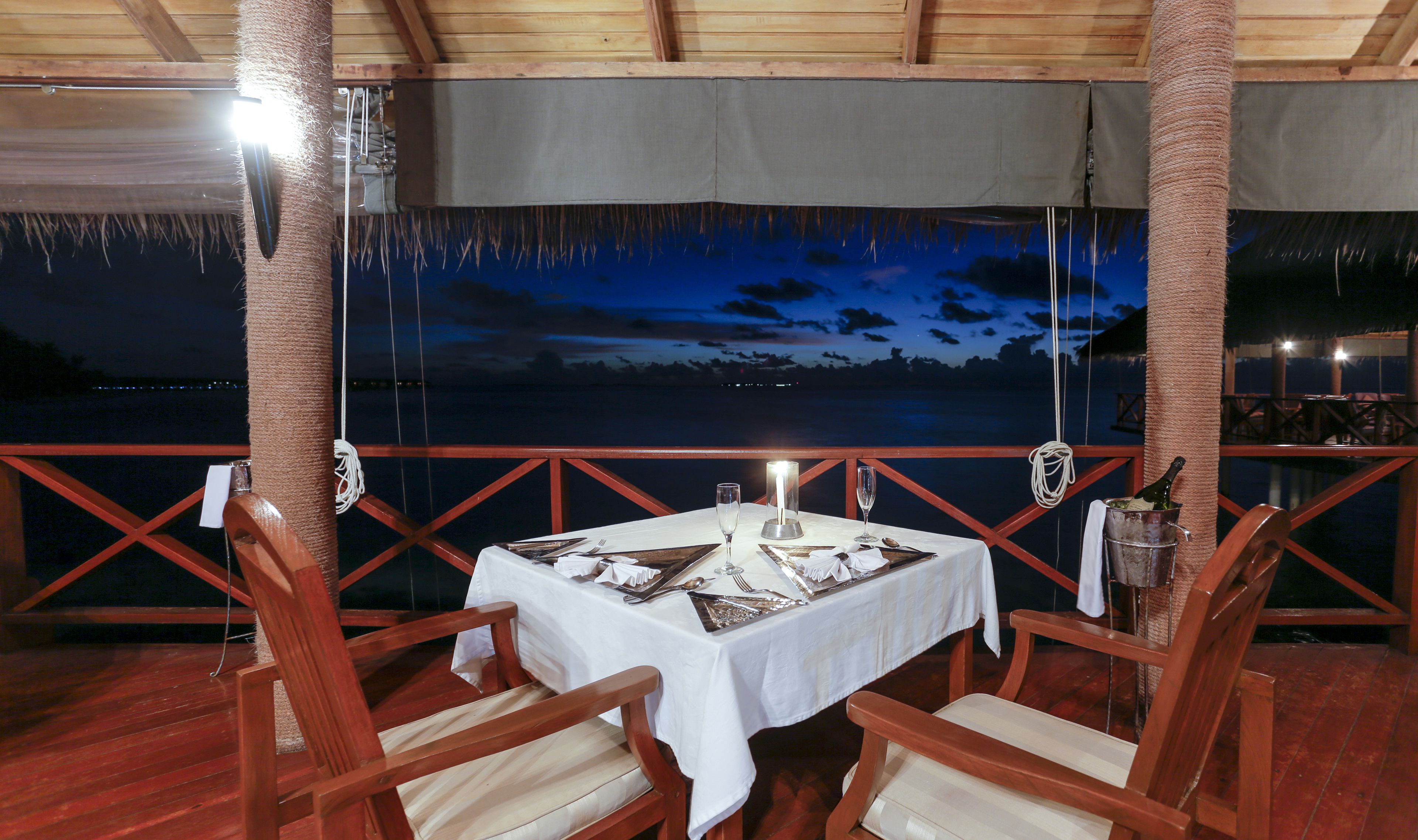 An outdoor dining area with a table set for two, overlooking a body of water at dusk.