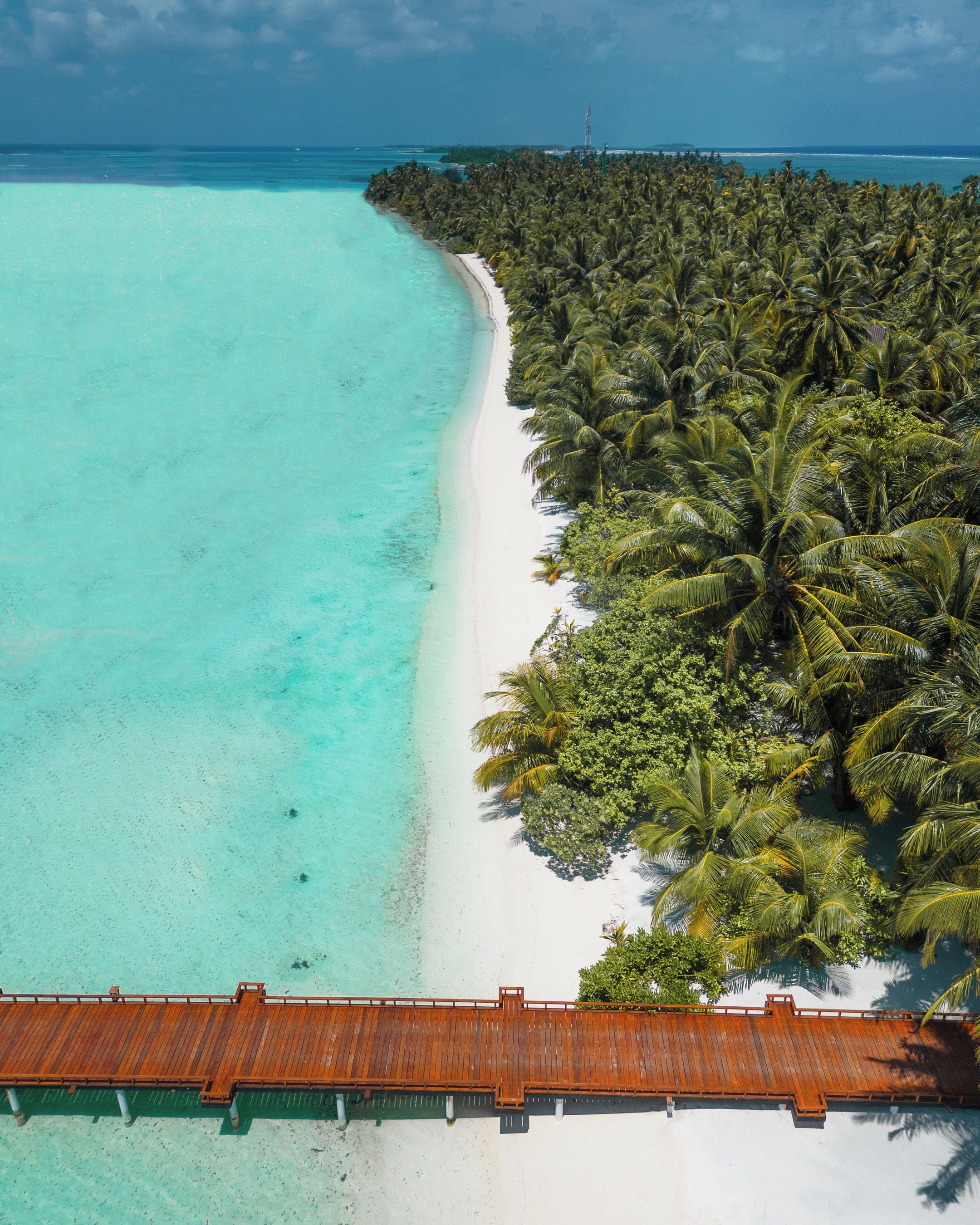 Ein Strand mit einer Holzpromenade, die zu einem weißen Sandstrand führt, umgeben von dichten Palmen und klarem türkisblauem Wasser.
