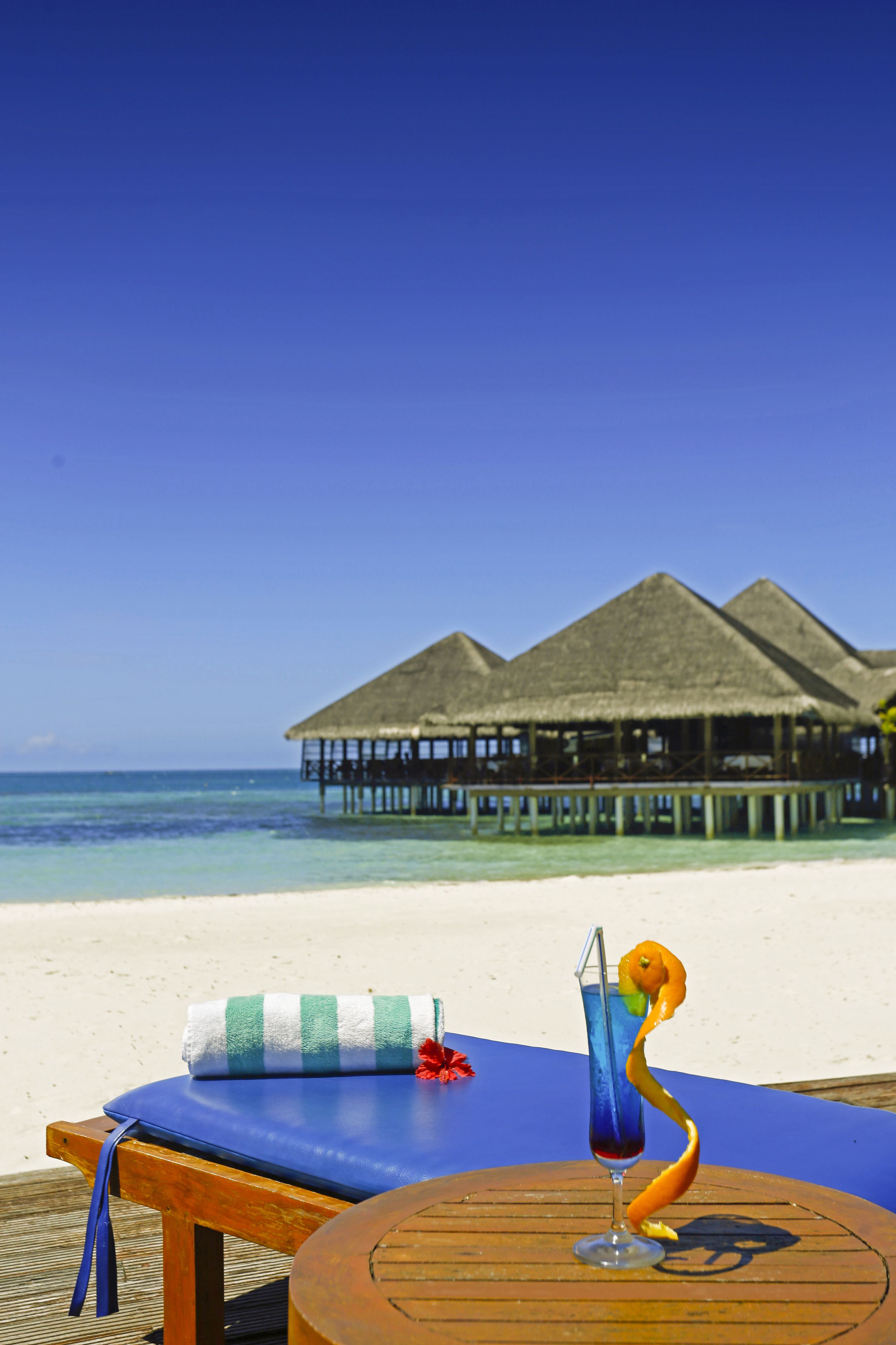 A beach view with overwater bungalows, a lounge chair with a towel, and a cocktail with a decorative fruit.