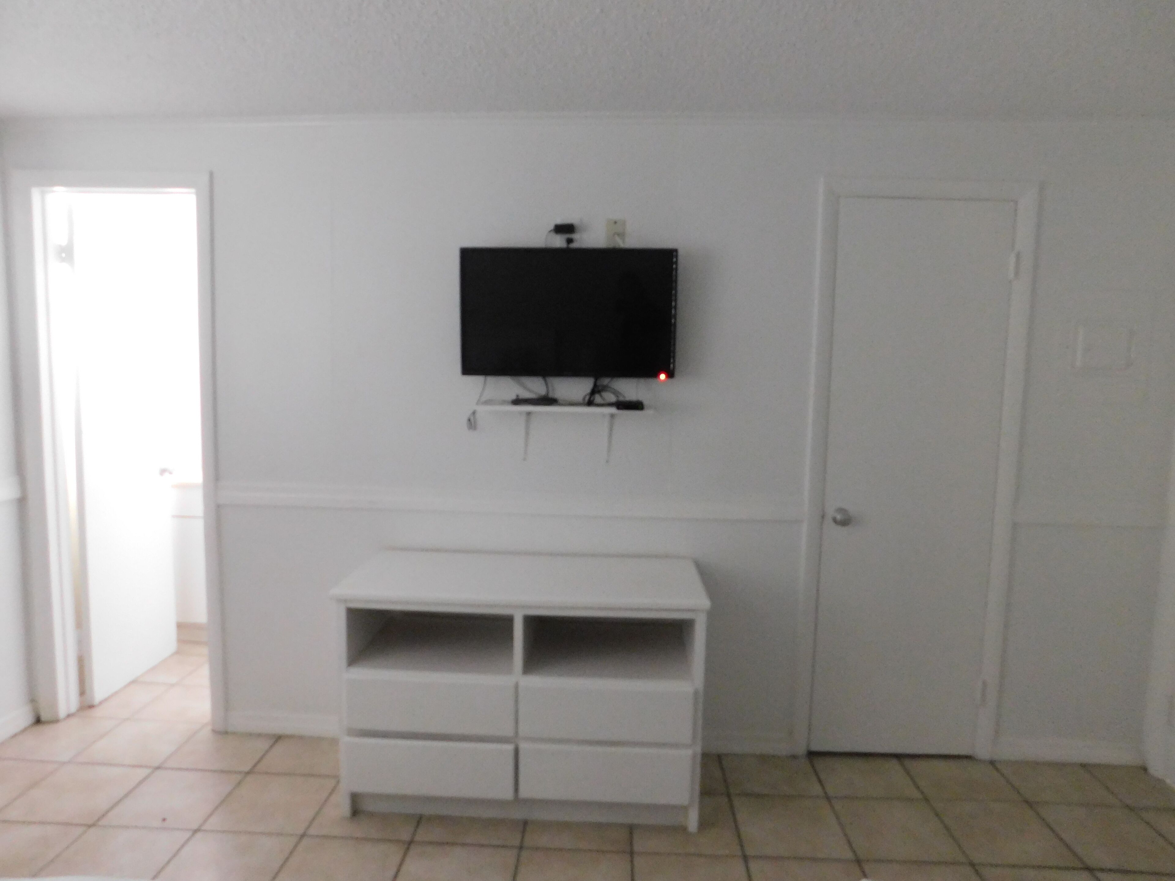 A room with a white wall-mounted TV, a white entertainment unit below it, and a closed door.