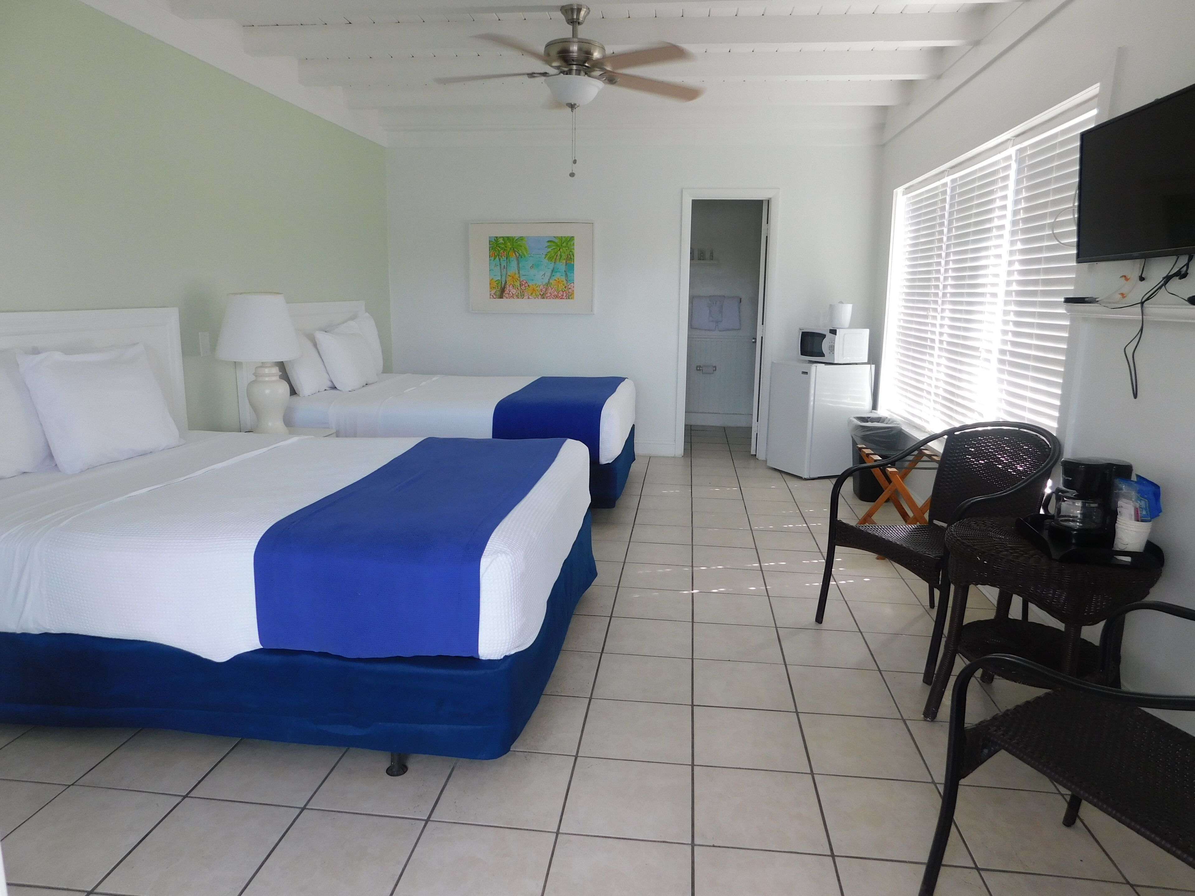 A hotel room with two beds, a ceiling fan, a TV, a small kitchenette, and a window with blinds.