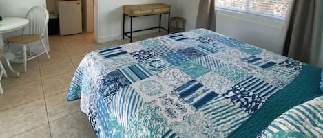 Une chambre à coucher comprenant un lit, un ventilateur de plafond, une télévision, une petite table, une chaise et une fenêtre avec des rideaux.