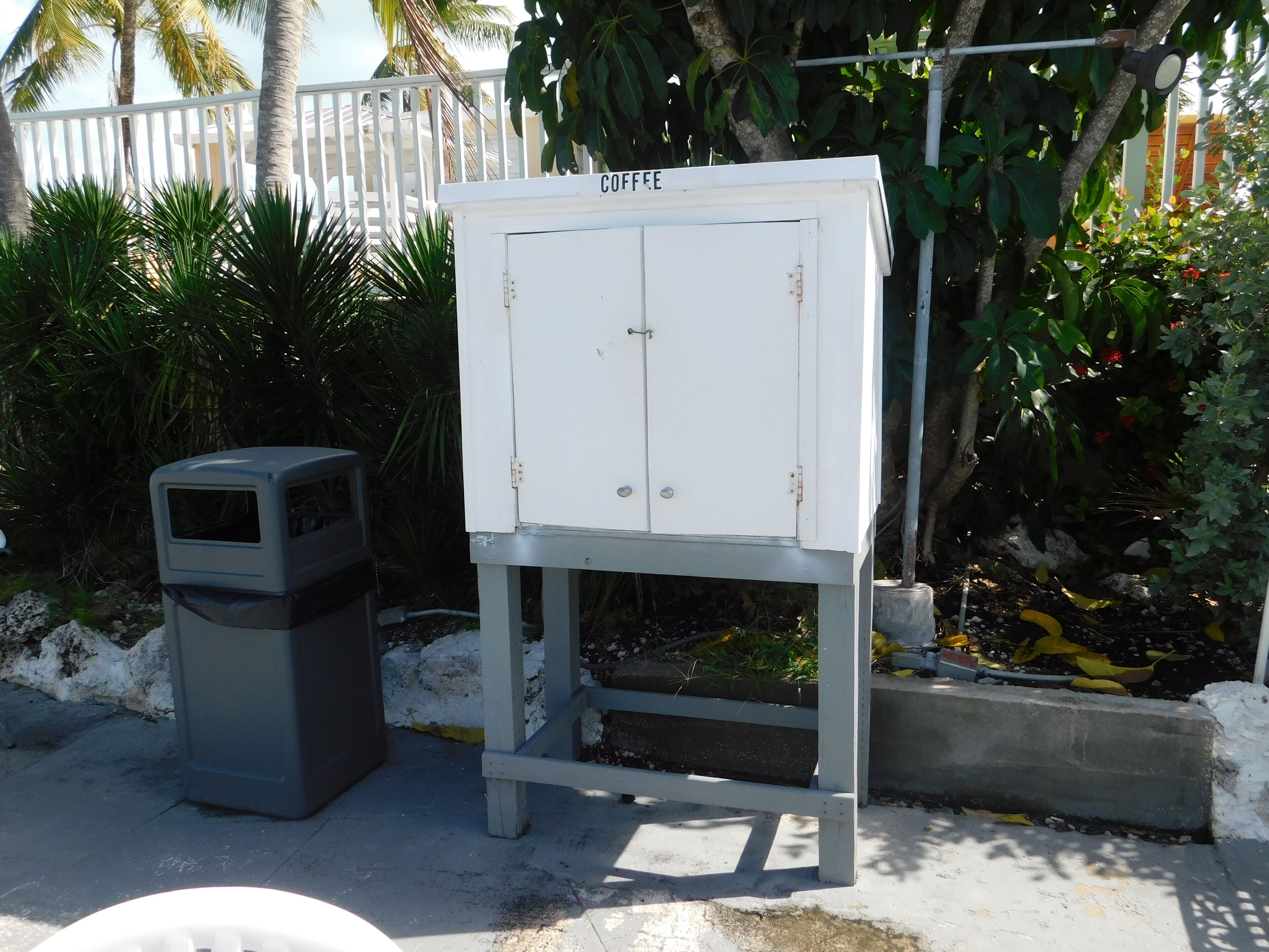 A white electrical box labeled "COFFEE" on a metal stand, a gray trash bin, and a concrete planter with yellow leaves.