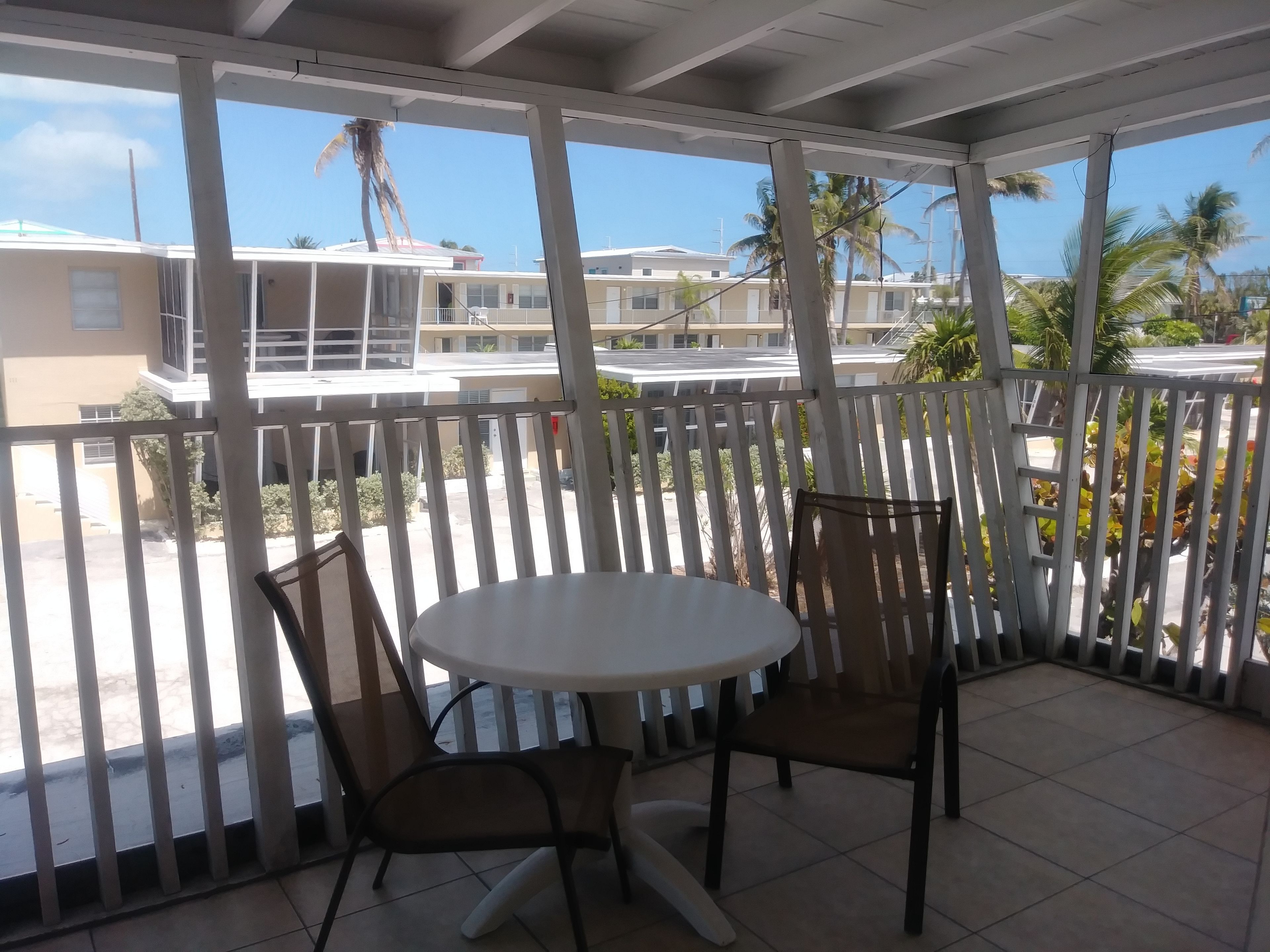 A covered outdoor area with a table and two chairs, overlooking a residential area with palm trees and buildings.
