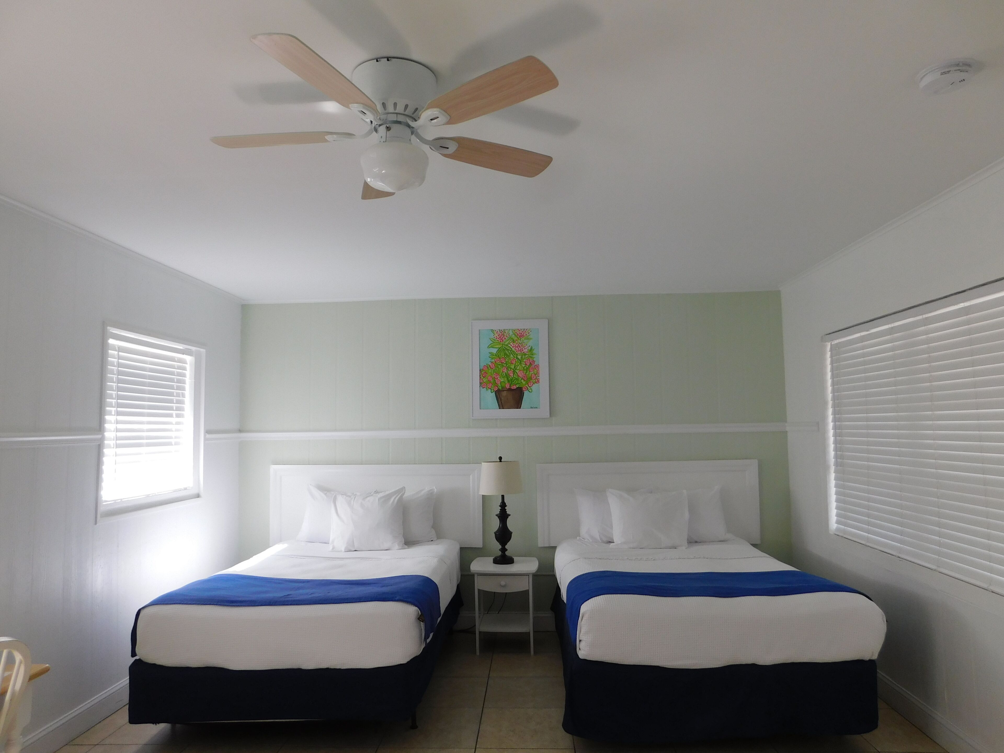 A room with two beds, a ceiling fan, a nightstand, a lamp, and a framed picture on the wall.