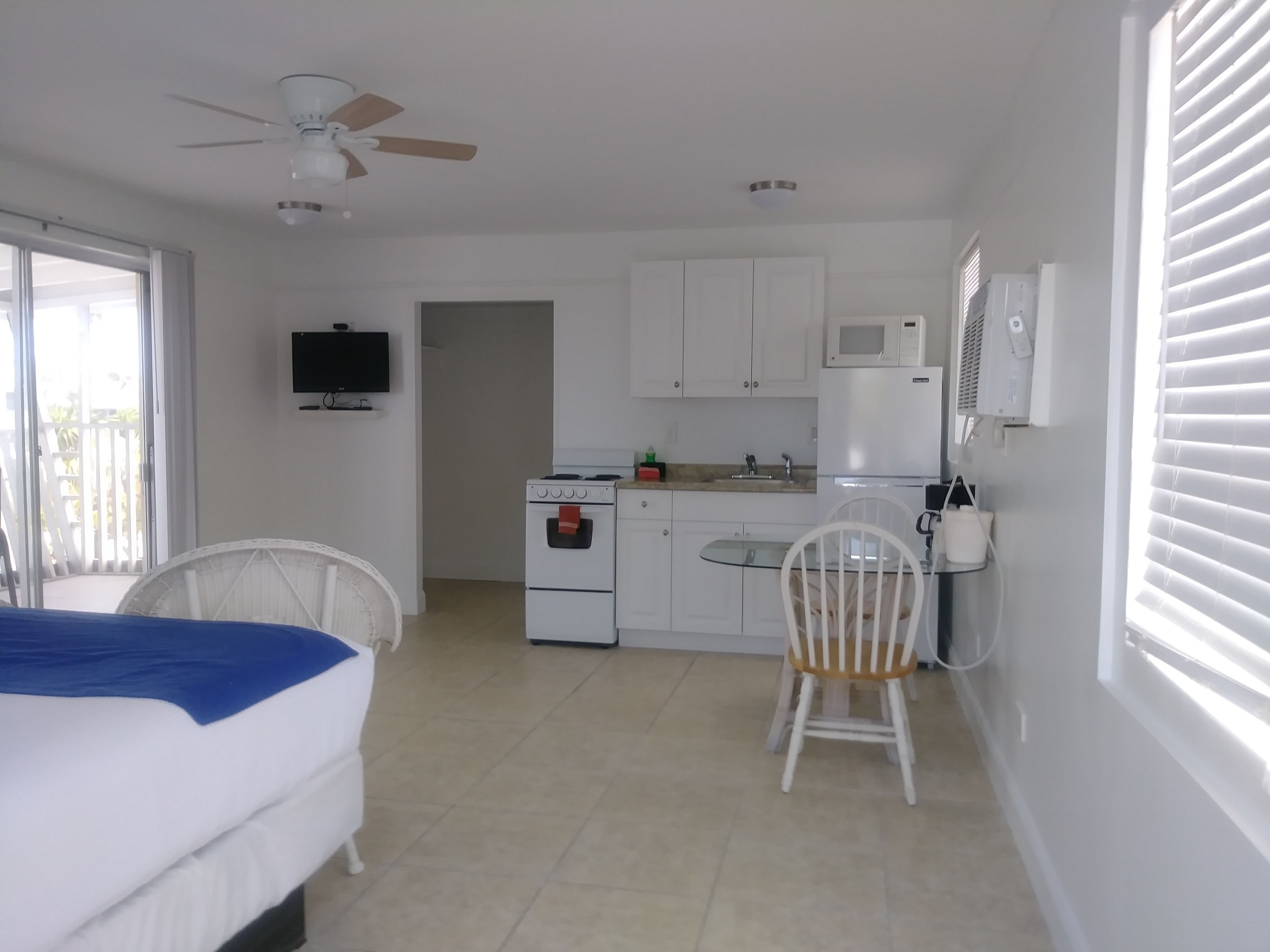 A compact room with a bed, a small kitchenette, a ceiling fan, and a TV.