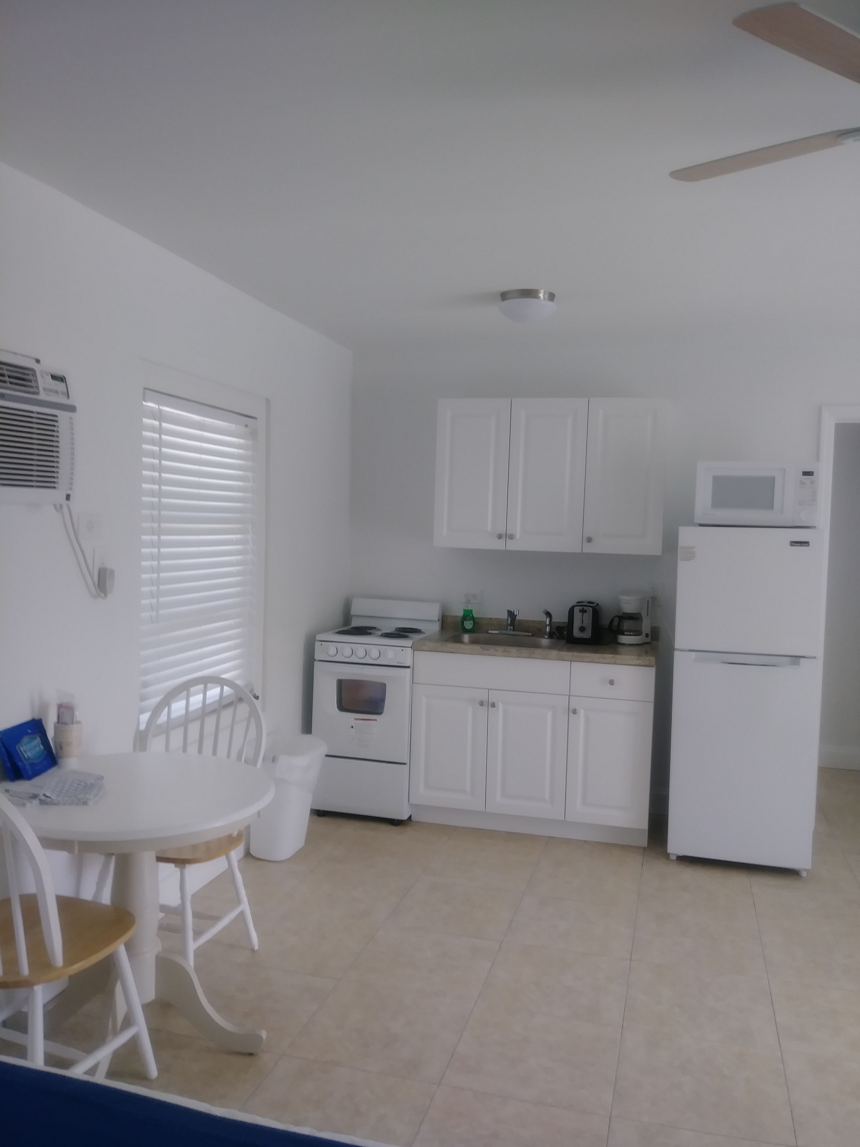 A compact kitchen with white cabinets, a stove, a microwave, and a refrigerator. There is a dining table with chairs and a window with blinds.