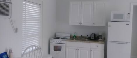 A compact kitchen with white cabinets, a stove, a microwave, and a refrigerator. There is a dining table with chairs and a window with blinds.