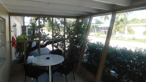 Une terrasse couverte avec une table ronde et des chaises, de grandes fenêtres et une vue sur la rue et la verdure.