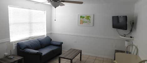 Una sala de estar con ventilador de techo, sofá, mesa de centro, televisor y un área de comedor con una silla y una mesa.