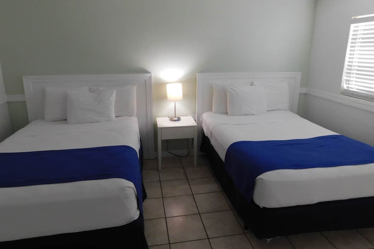 Ein Hotelzimmer mit zwei Betten, jeweils mit weißer Bettwäsche und blauen Akzenten, einem kleinen Nachttisch mit Lampe sowie einem Fenster mit Jalousien.
