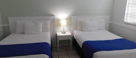A hotel room with two beds, each with white bedding and blue accents, a small bedside table with a lamp, and a window with blinds.