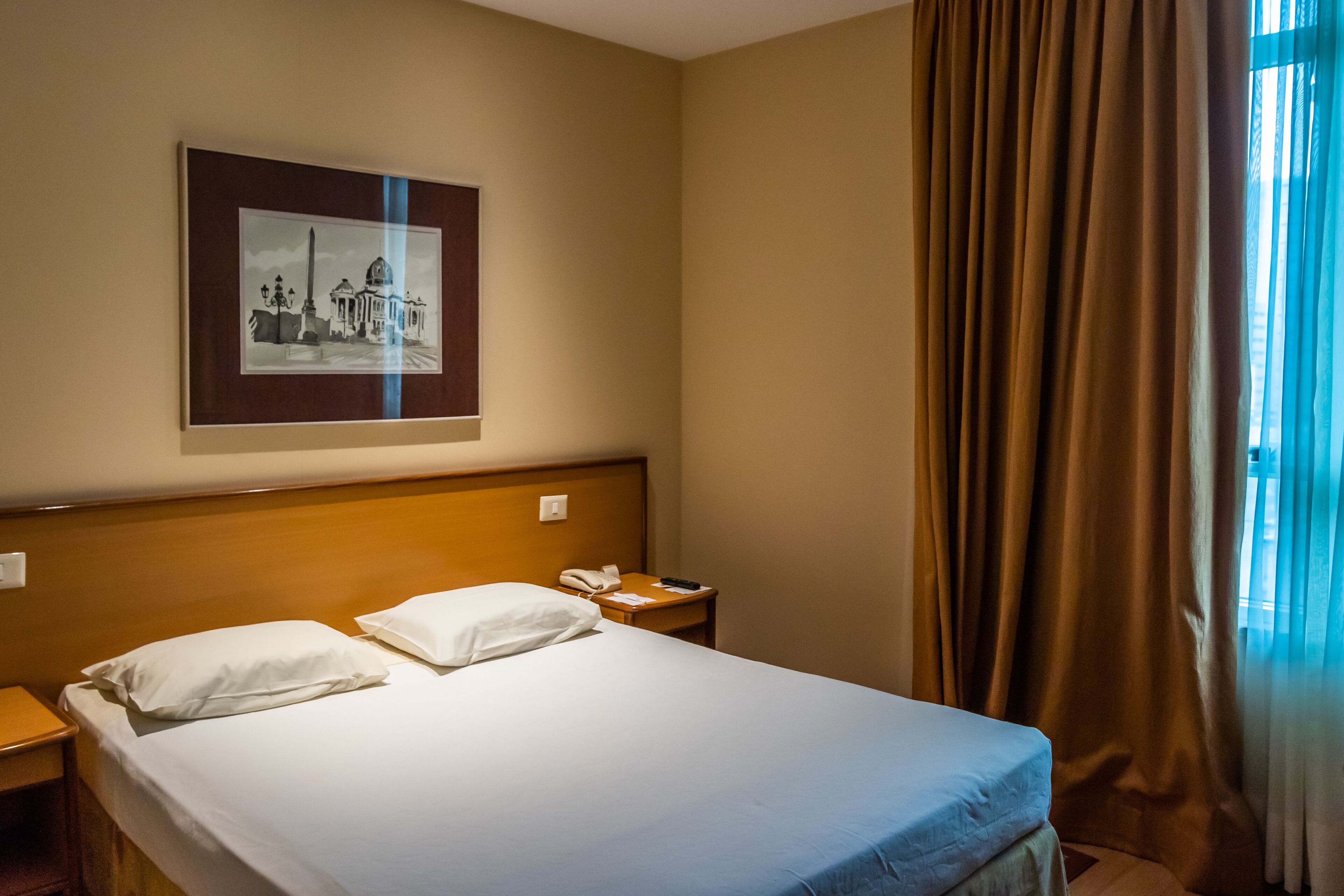 A hotel room with a bed, a nightstand, a framed picture on the wall, and a window with curtains.