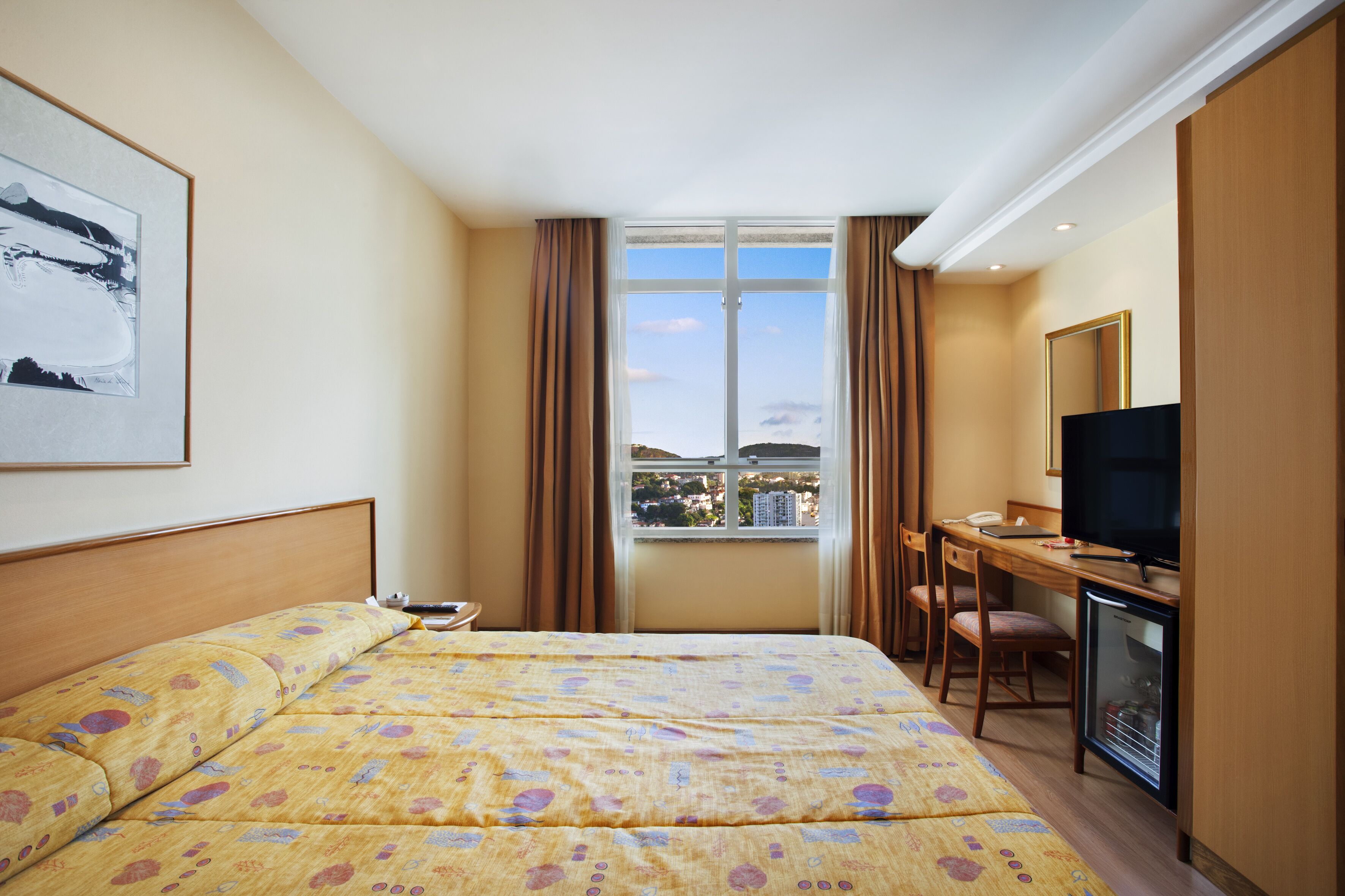 A hotel room with a bed, a desk with a television, a small refrigerator, and a window with curtains.