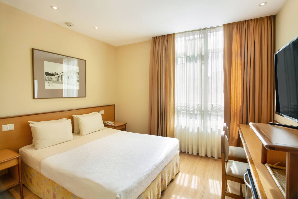A hotel room with a large bed, a desk with a chair, a television, and a window with curtains.