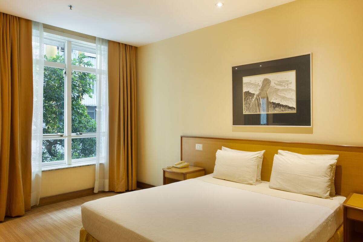 A hotel room with a large bed, a bedside table with a phone, a framed picture on the wall, and a window with curtains.