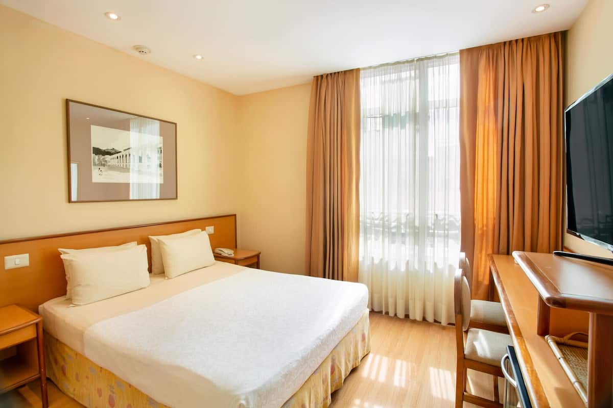A hotel room with a bed, bedside tables, a desk, a chair, a TV, and a window with curtains.