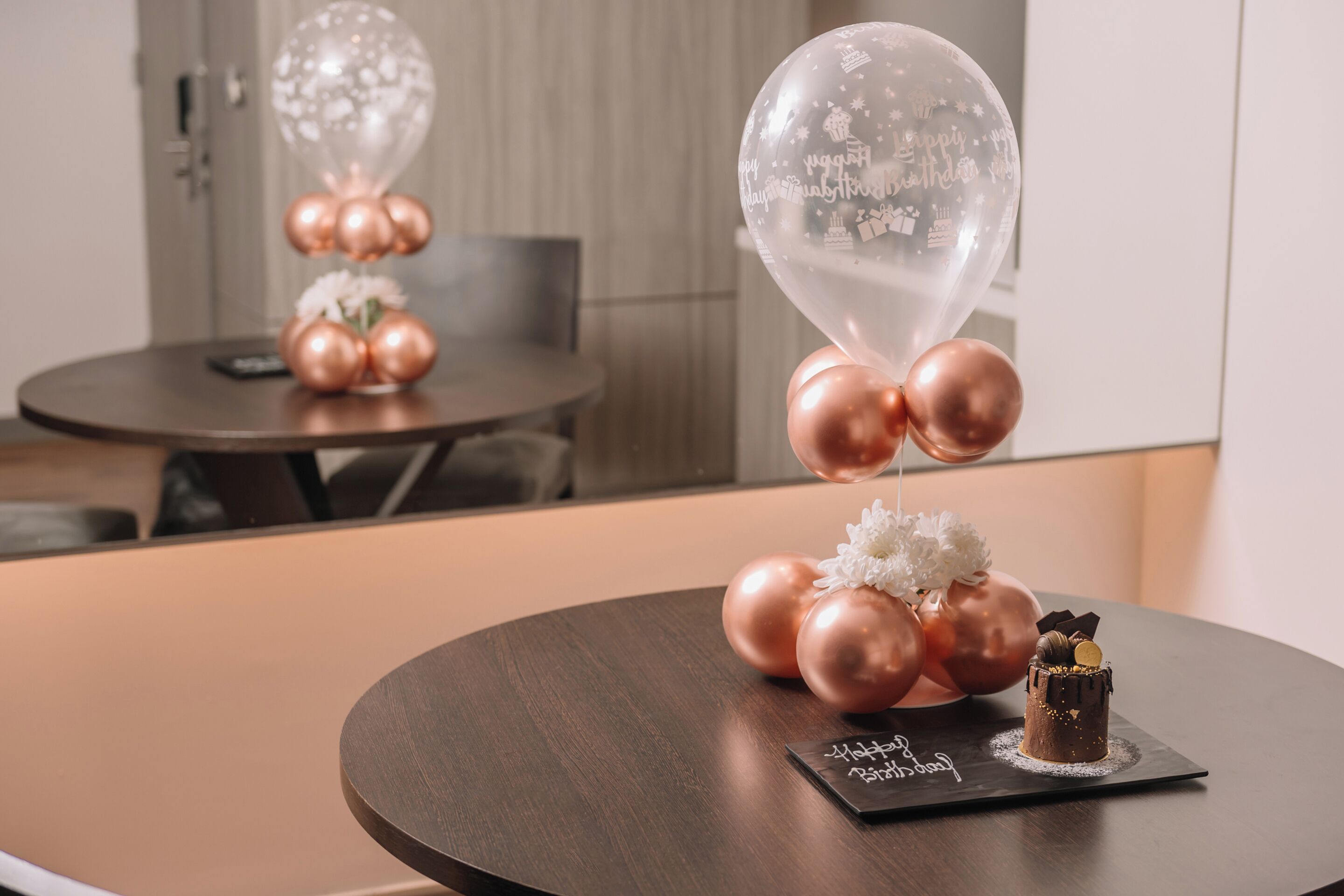 A round table with a small chocolate cake and a card, surrounded by balloons and flowers.