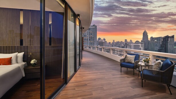 Un balcón moderno con vistas a un horizonte urbano al atardecer, que incluye un sofá, sillas y una pequeña mesa con flores.