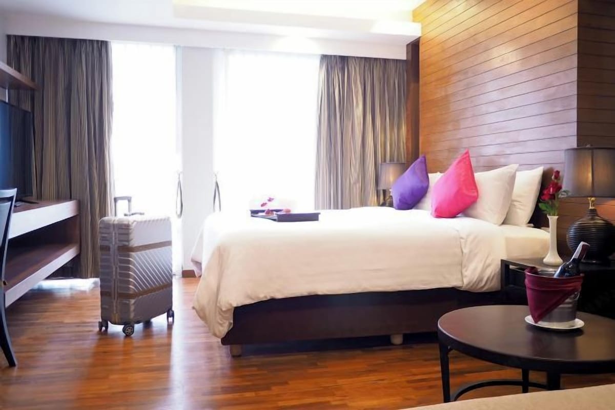 A hotel room with a bed, a desk, a suitcase, a table with a bottle, and a lamp.