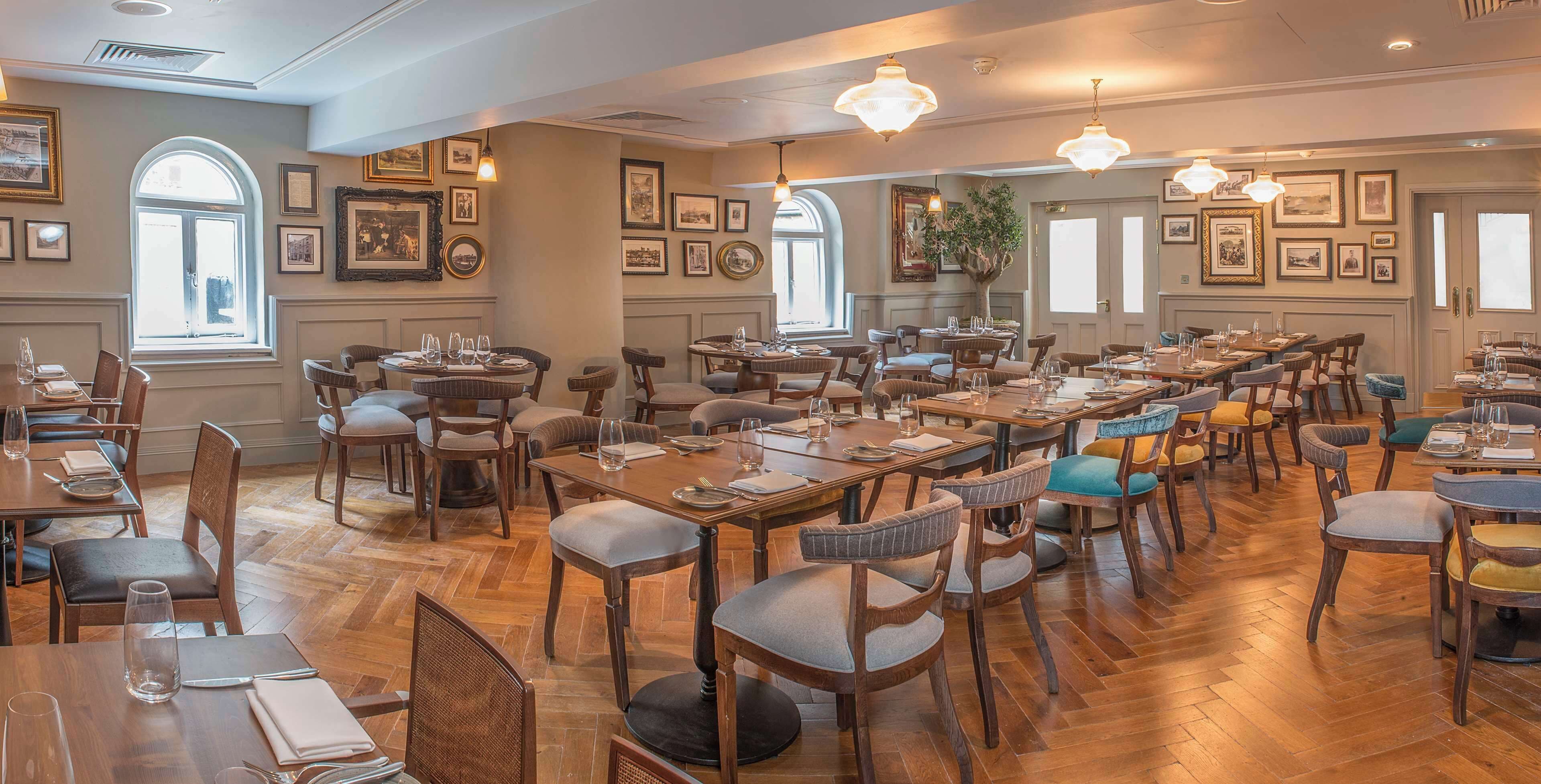 Un restaurante amplio con varias mesas y sillas, cuadros en las paredes y ventanas grandes que dejan entrar luz natural.