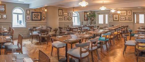 A spacious restaurant with multiple dining tables and chairs, framed pictures on the walls, and large windows providing natural light.