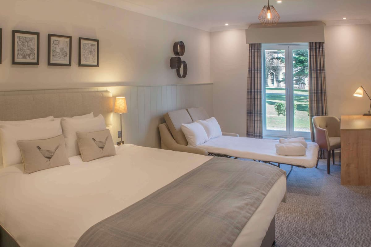 A hotel room with a large bed, a smaller bed, a desk, and a chair. There are framed pictures on the wall and a window with a view of greenery.