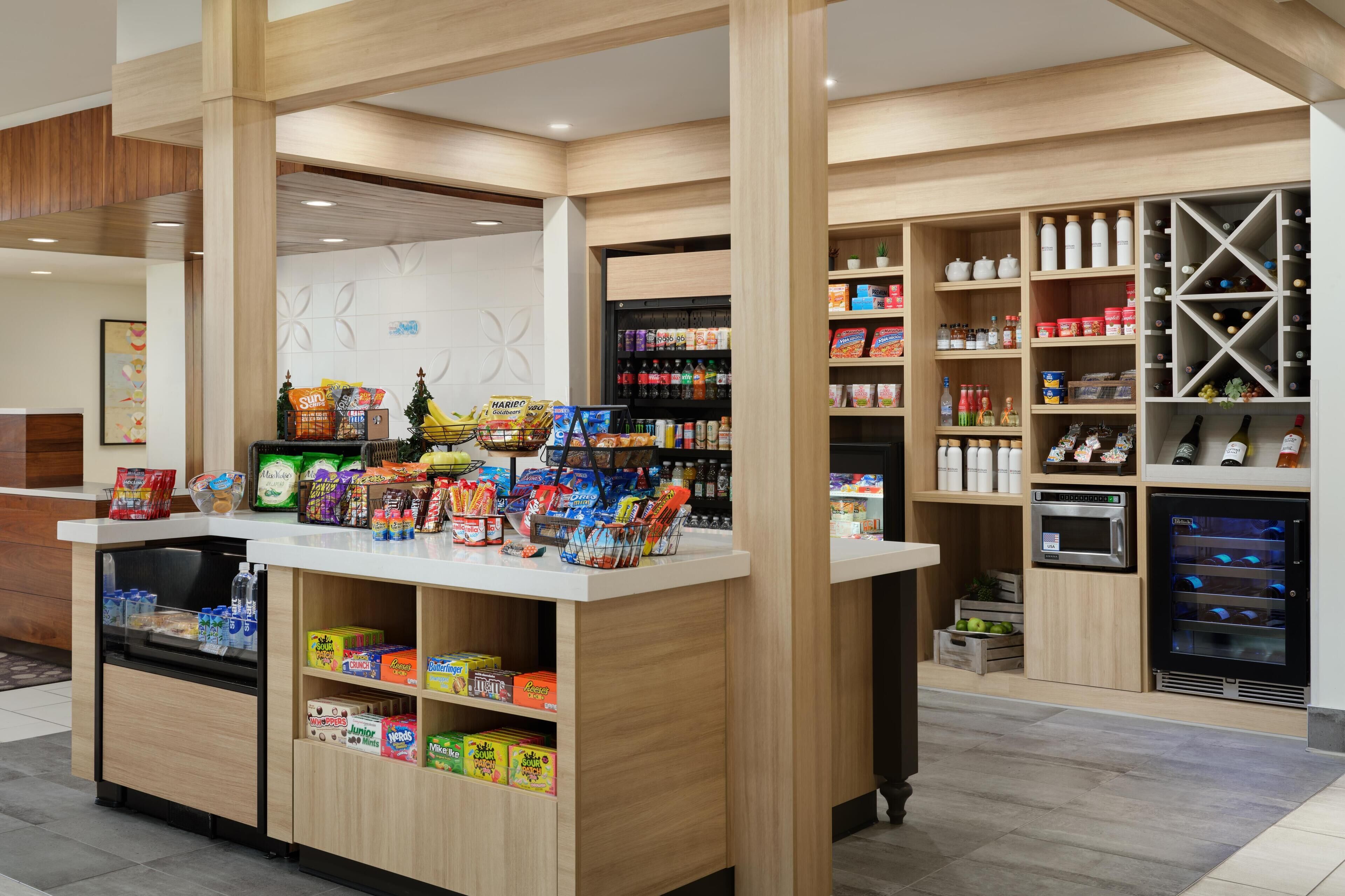 Un supermercado moderno con un mostrador central repleto de bocadillos y bebidas, estantes con agua embotellada y snacks, y un refrigerador de vinos al fondo.