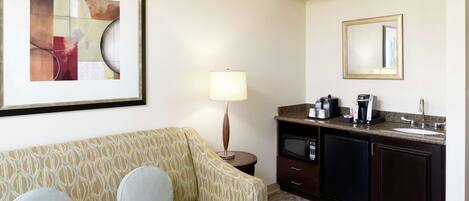 A hotel room with a patterned sofa, a small round table with a lamp, a kitchenette with a microwave, and a framed abstract painting on the wall.