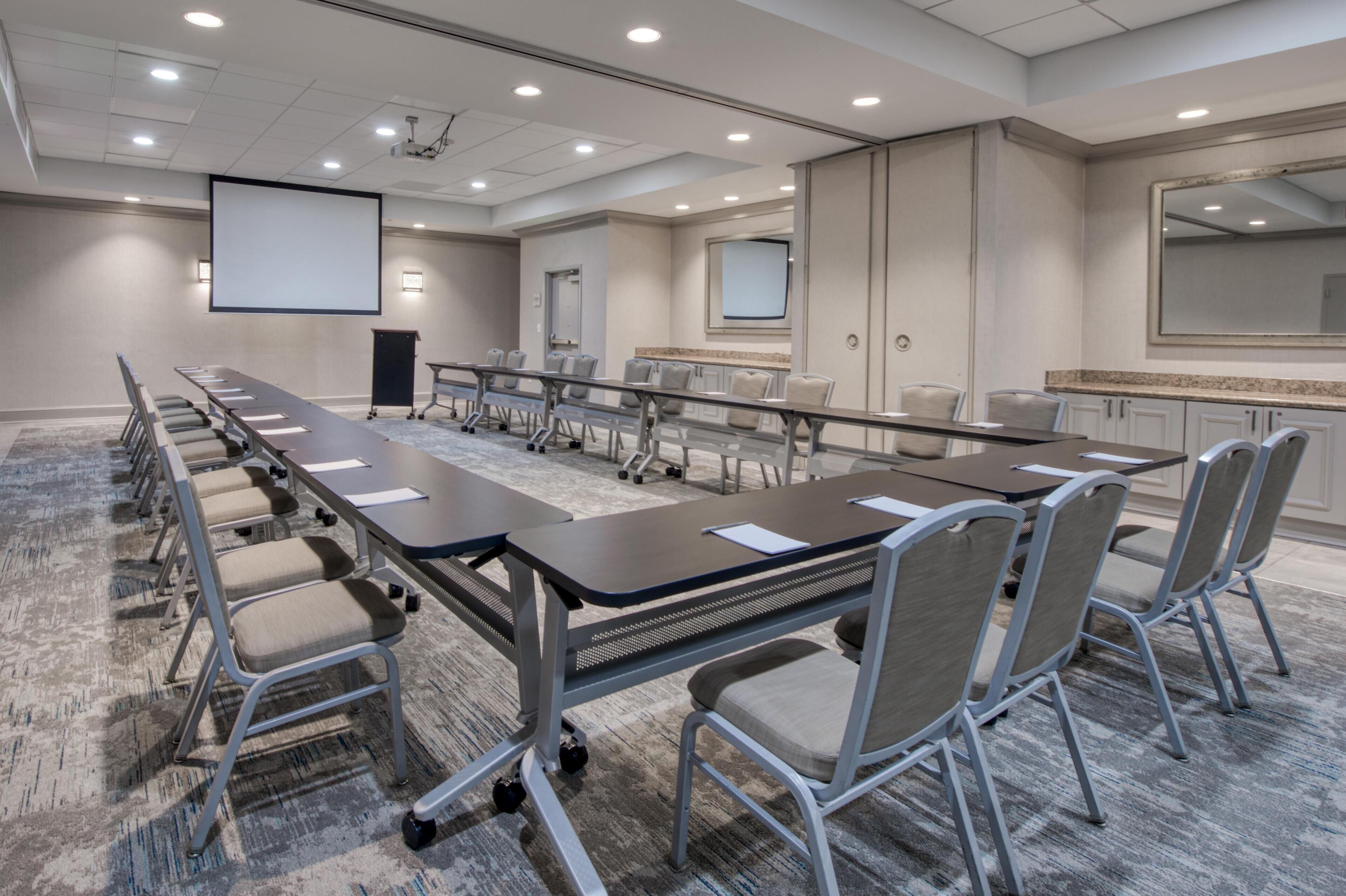 Un salón de conferencias con una larga mesa, sillas, una pantalla de proyector y un espejo grande.