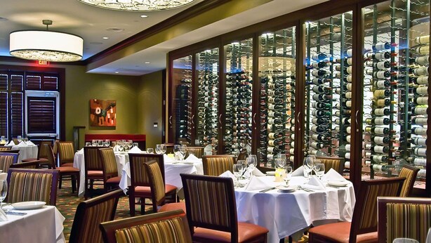 A restaurant with tables set for dining, a wine wall, and a chandelier. - Hilton Garden Inn Durham Southpoint (Durham)