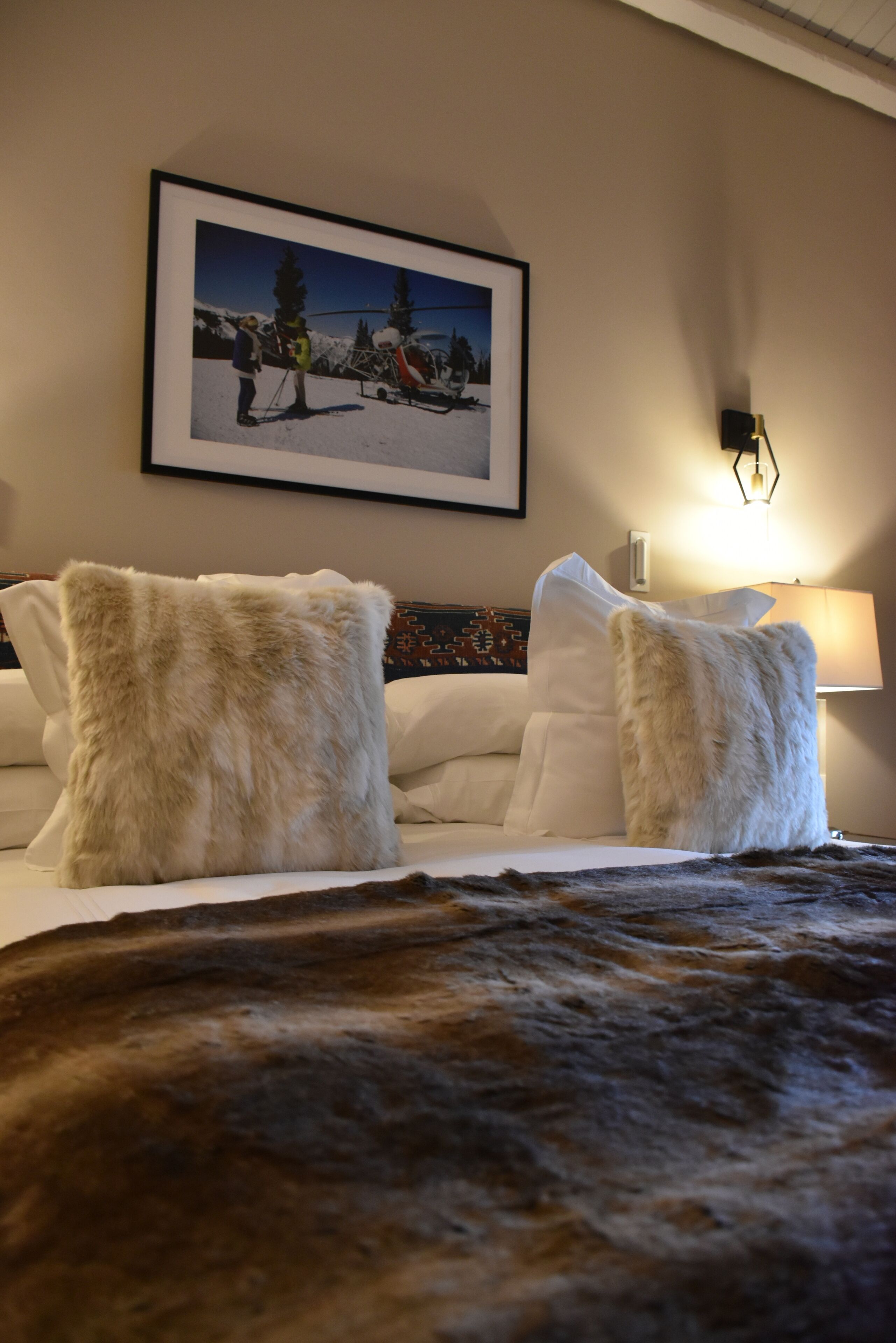 A hotel room with a bed, two white pillows, a framed picture of a snowy landscape, and a wall-mounted lamp.