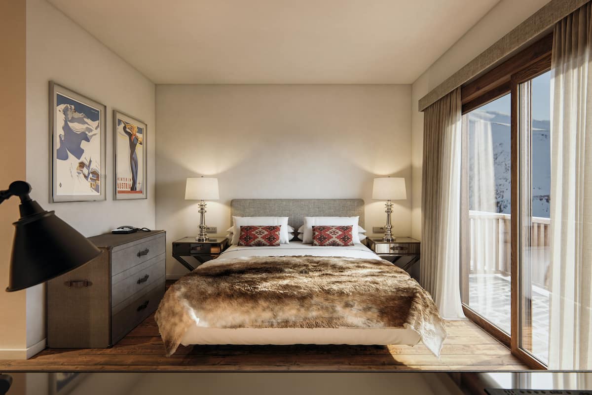 Habitación de hotel con una cama grande, dos mesitas de noche con lámparas, un aparador y una ventana con vista a un balcón.