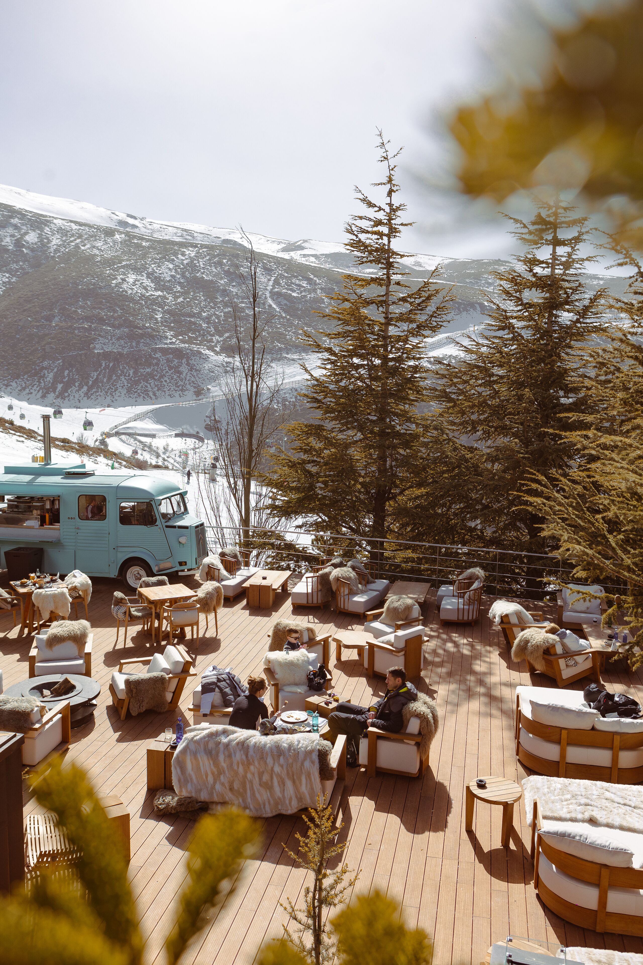 A wooden deck with outdoor seating and a vintage van, surrounded by snow-covered mountains and trees.