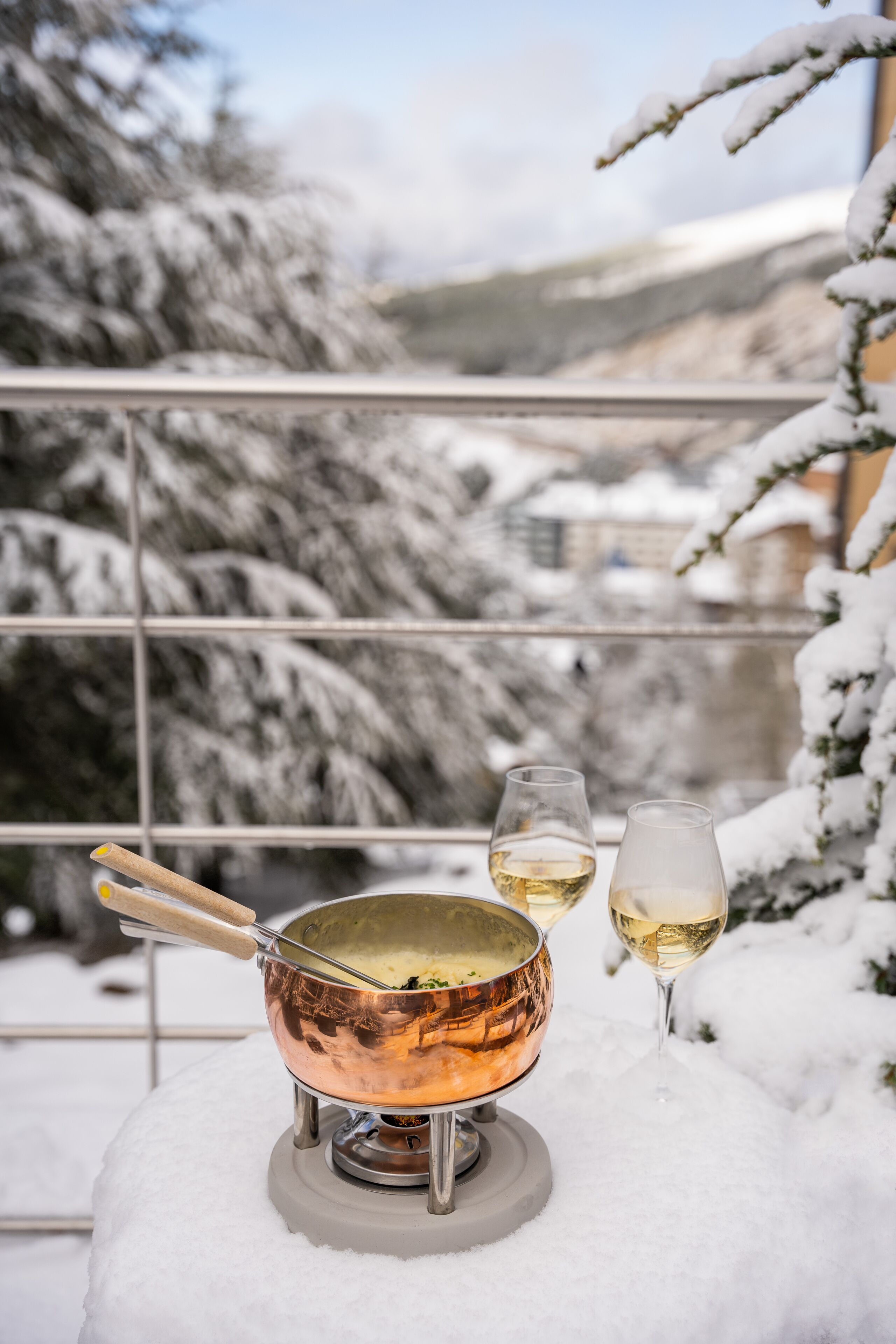 A copper pot on a portable stove with two glasses of white wine on a snowy balcony.