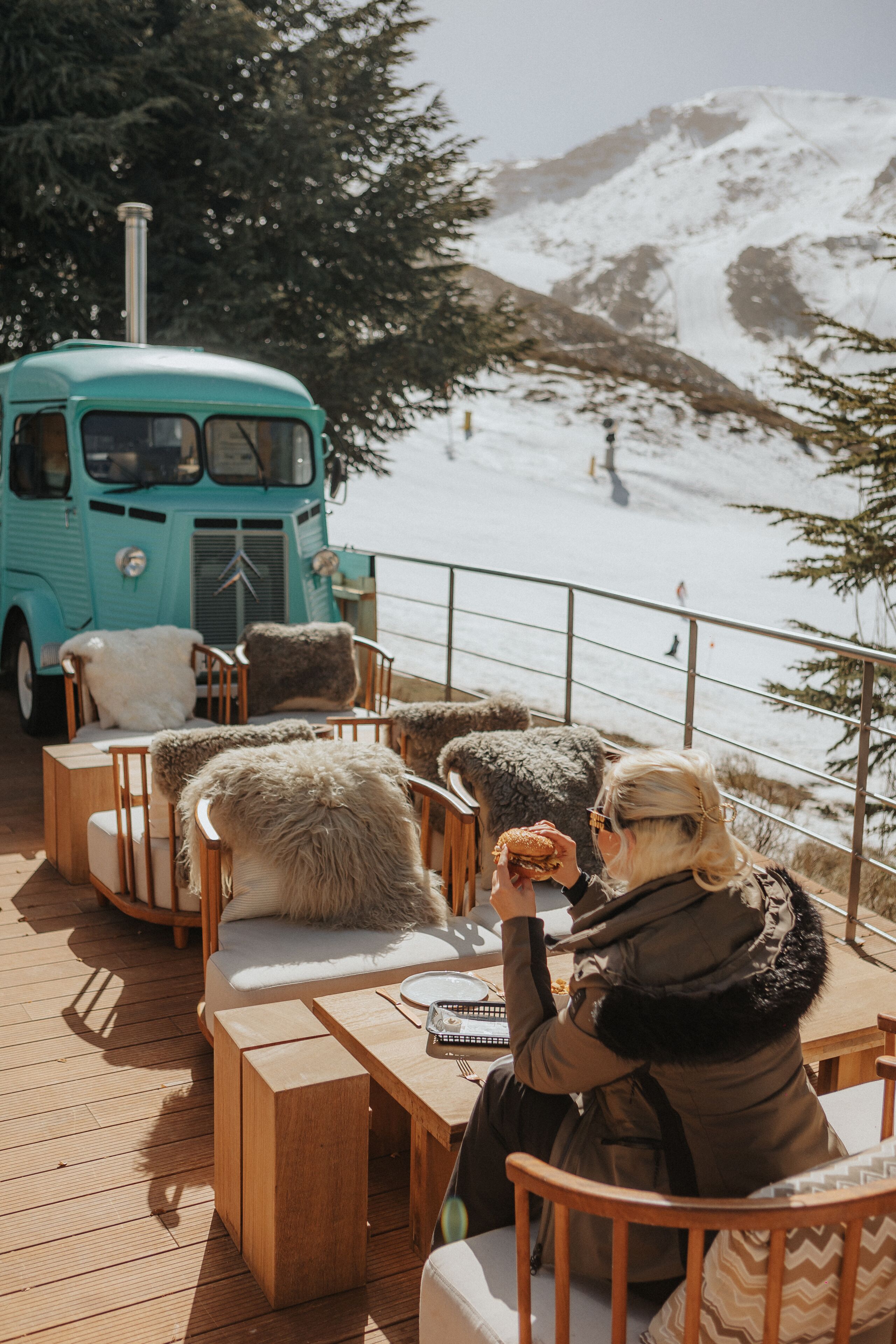 A wooden deck with outdoor seating, a turquoise vintage bus, and a snowy mountain backdrop.