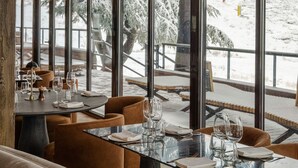 A restaurant with a glass table and brown upholstered chairs, large windows with a snowy view, and a wooden ceiling.