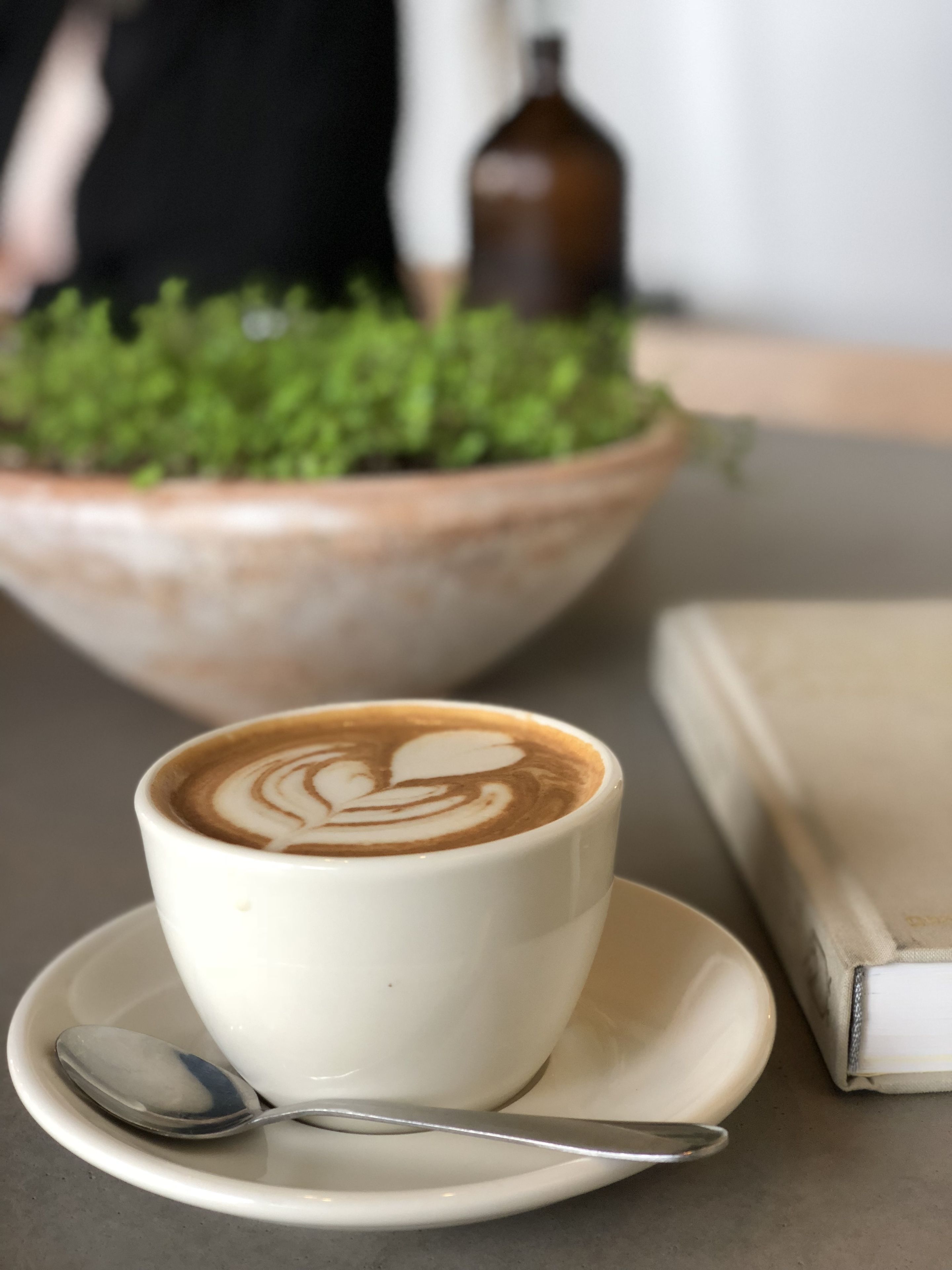 Une tasse de café ornée d’un motif en latte art, posée sur une soucoupe, avec une cuillère et un livre sur la table.