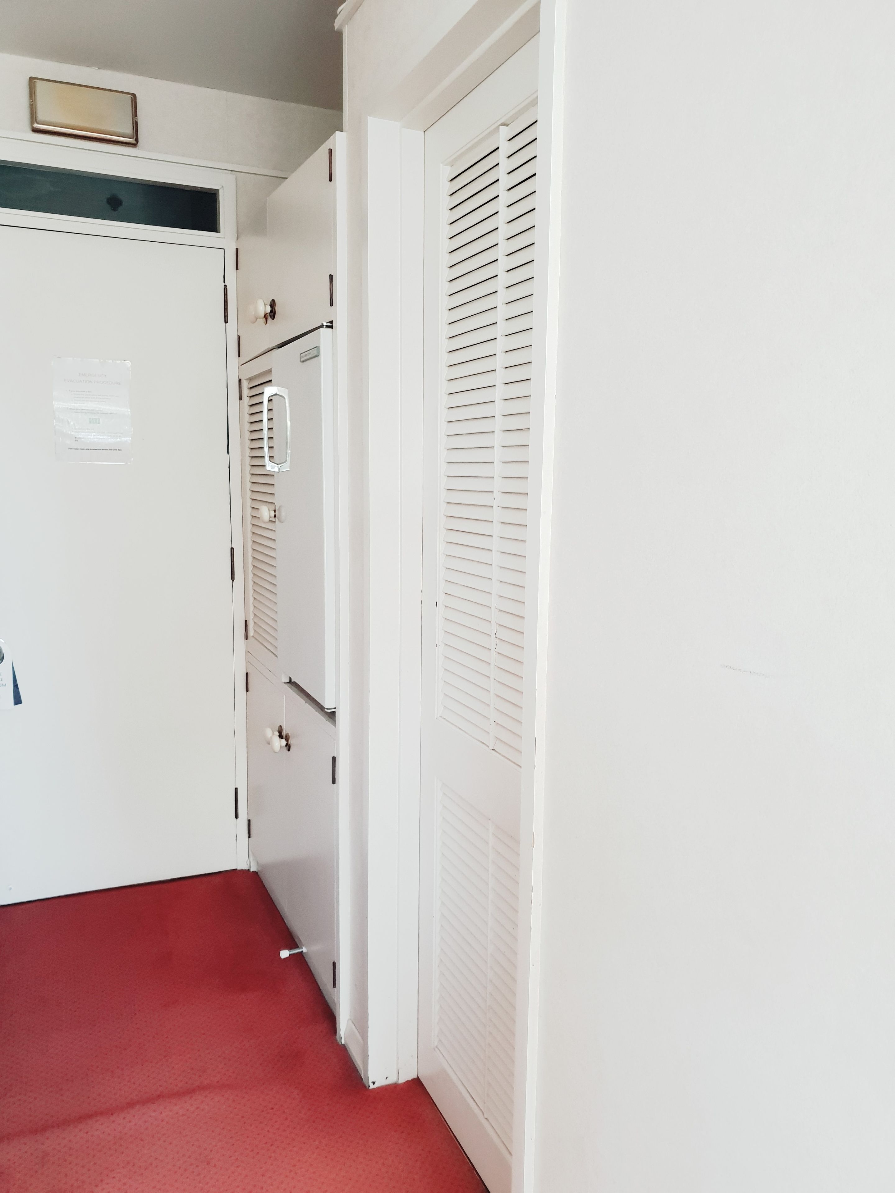 Un couloir avec des portes blanches, une moquette rouge et une grille de ventilation au mur.