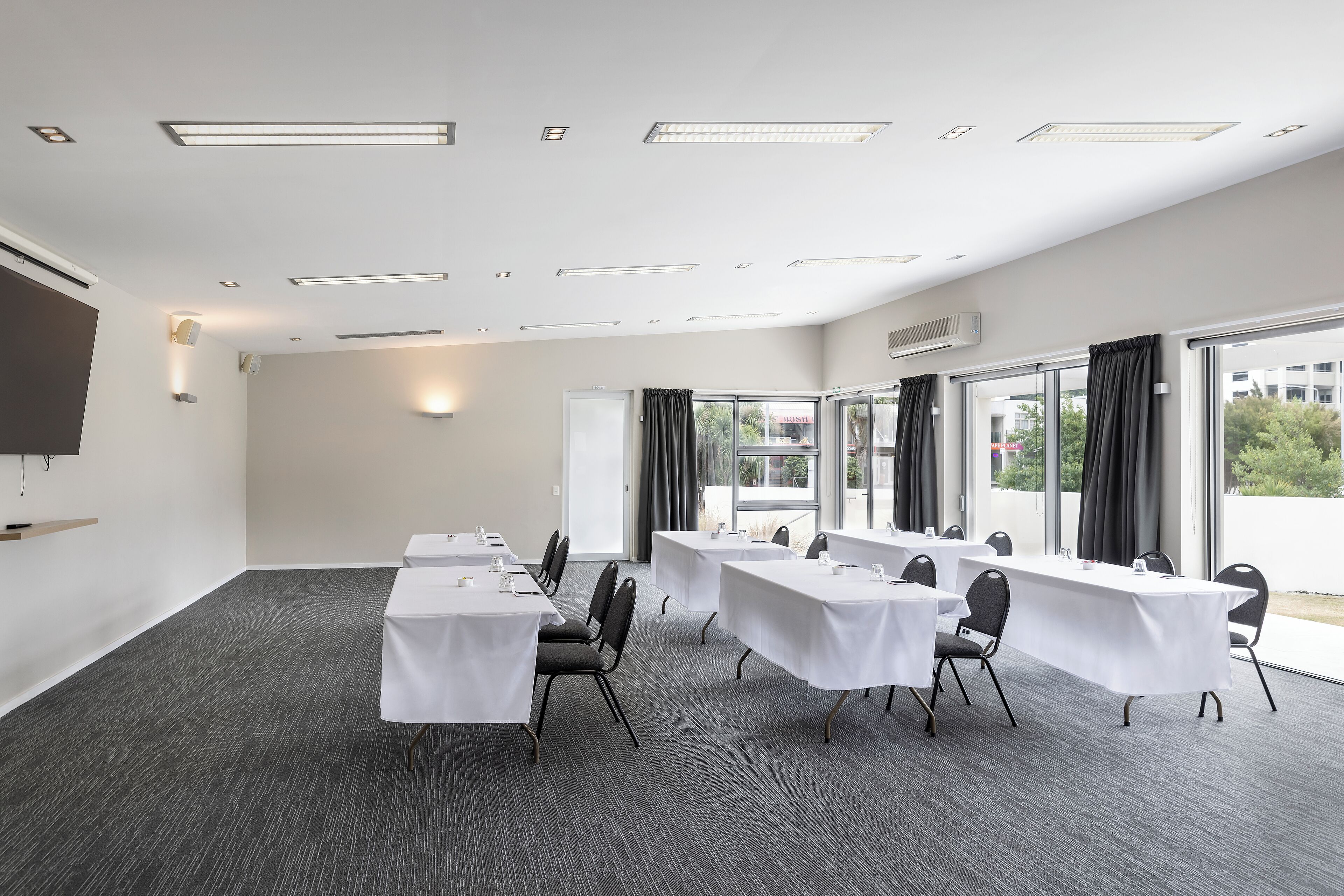 Une salle de conférence équipée de tables et de chaises, de grandes fenêtres avec des rideaux et d’un éclairage au plafond.