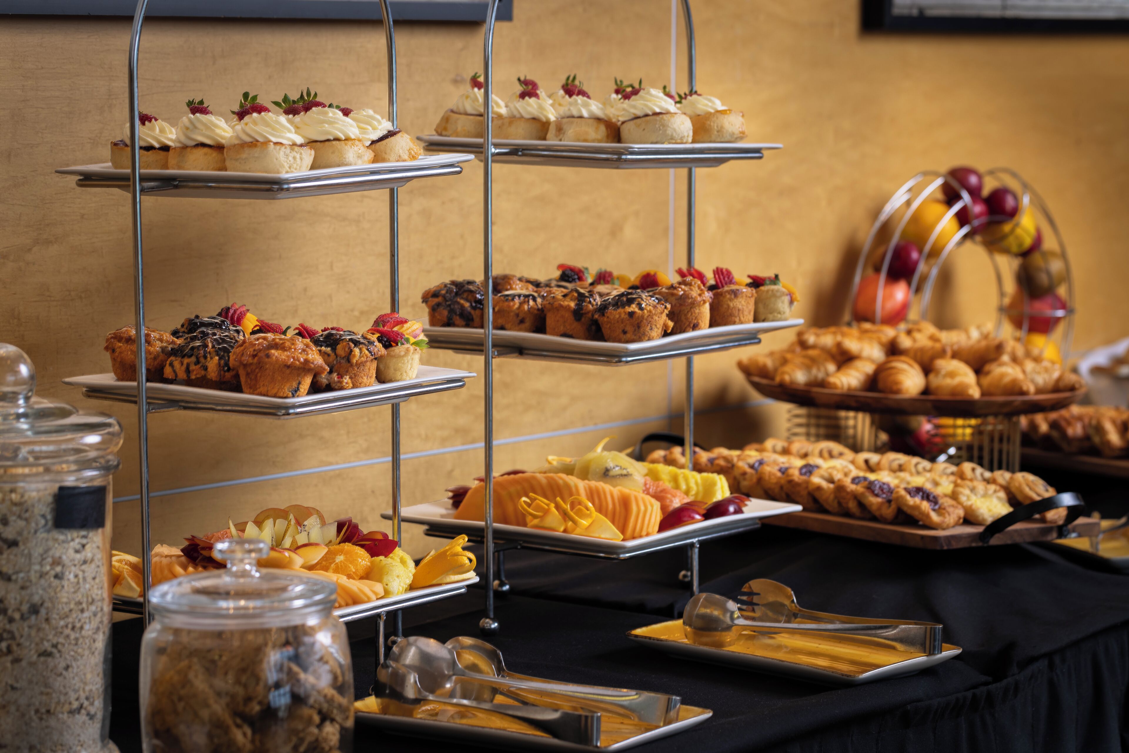 Un bufé con múltiples bandejas de pasteles, muffins y fruta.